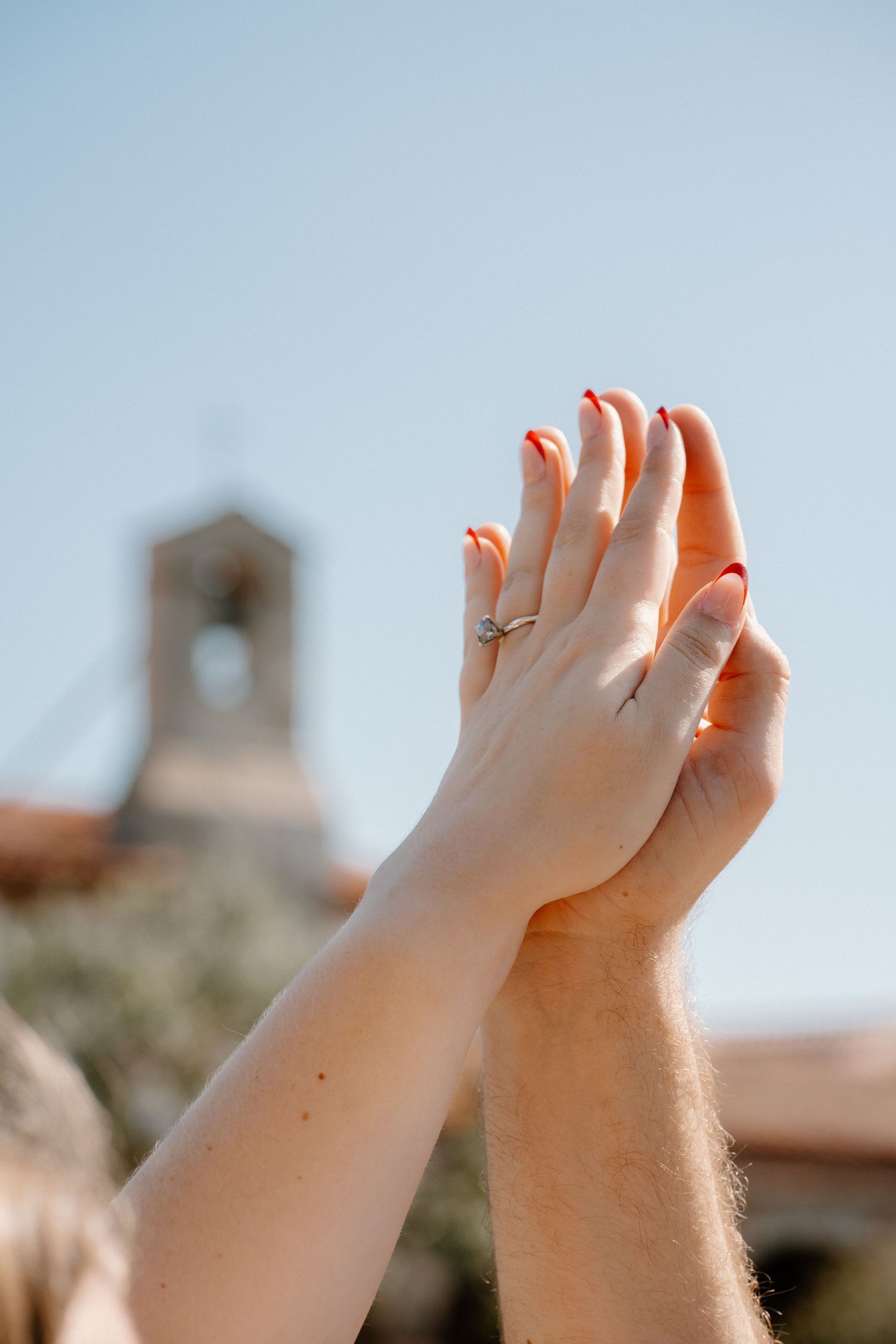 Two hands raised together showing an engagement ring with diamond on finger, red nails, church bell tower in background.