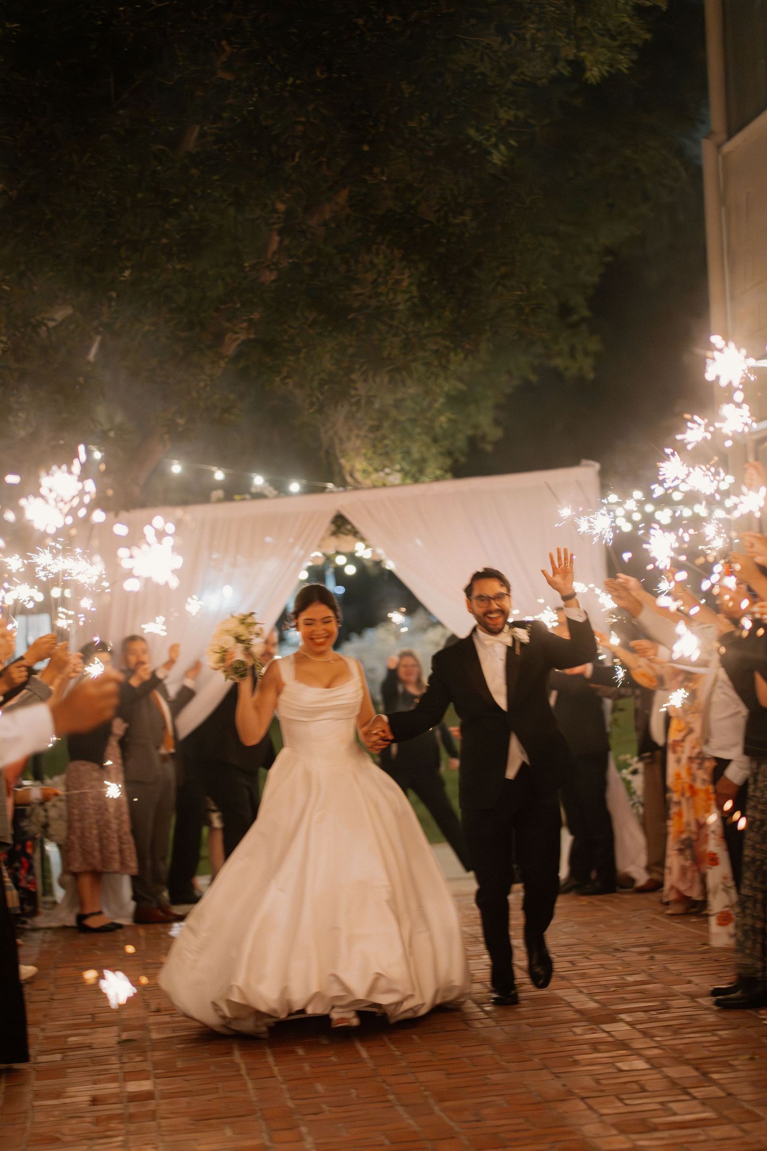 Bride and groom exit wedding reception through sparkler tunnel at night, waving joyfully to guests.