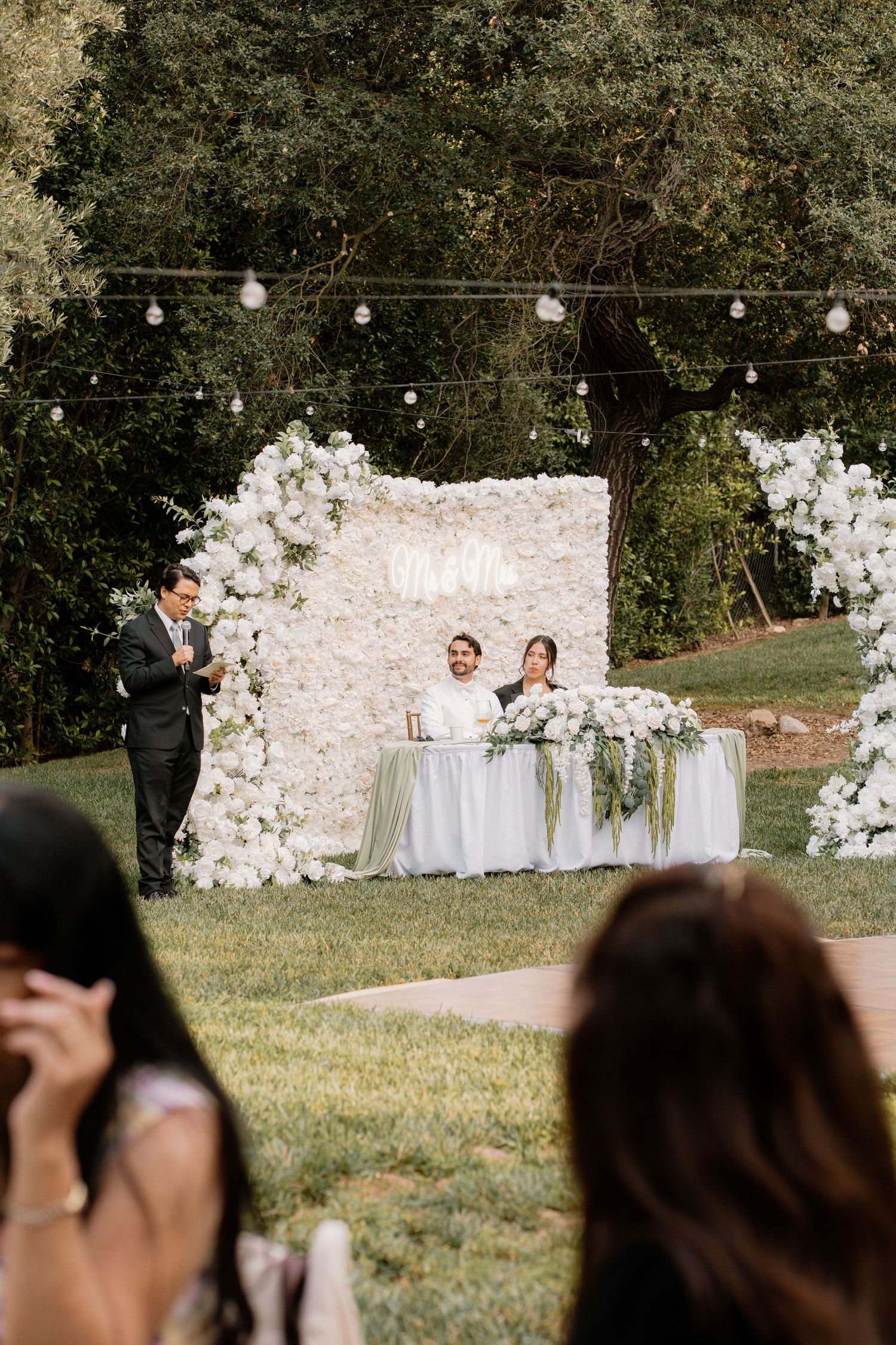 Outdoor wedding ceremony with couple at floral white flower wall backdrop, string lights, and lush greenery.