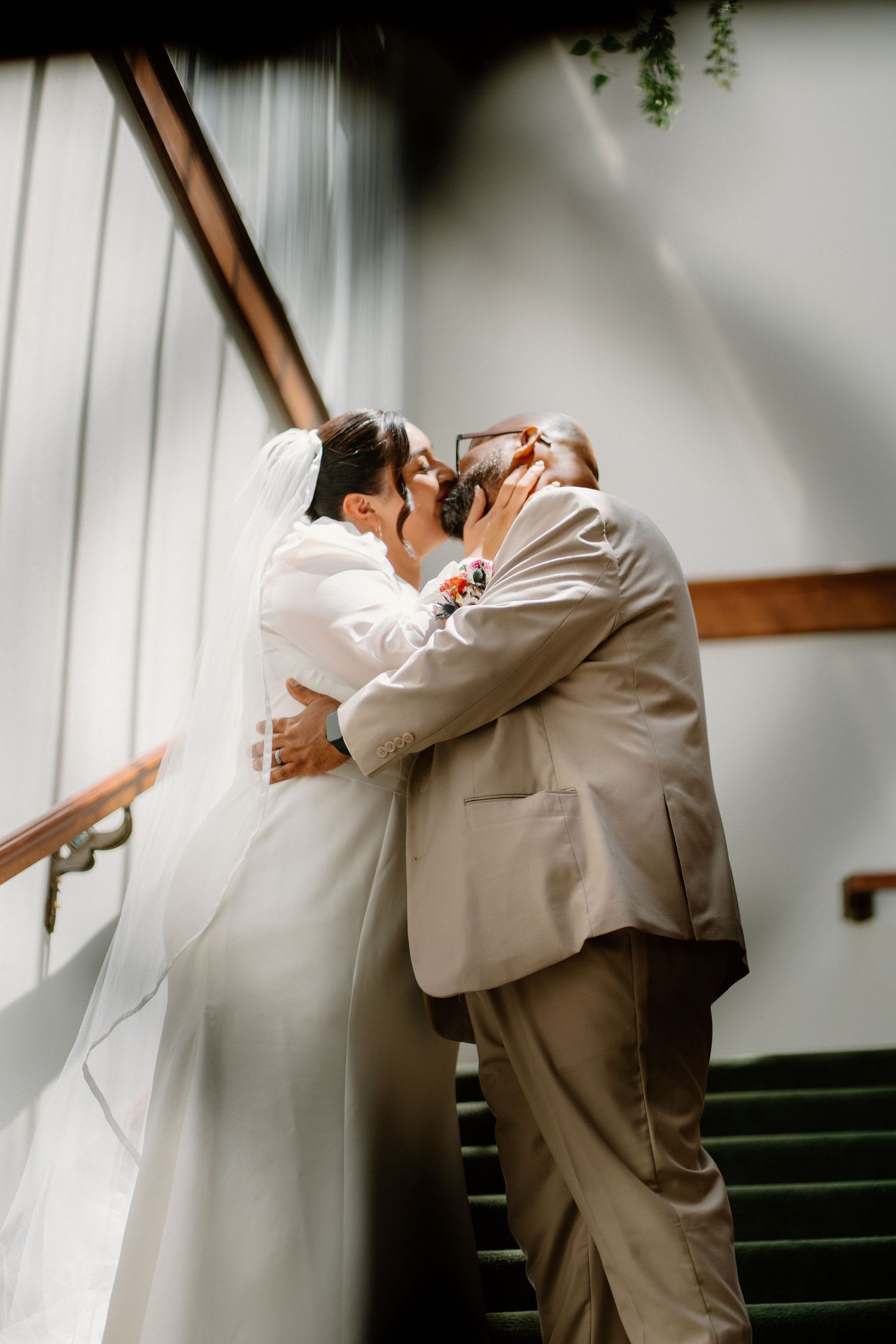 Bride and groom share a romantic kiss on a staircase, embracing in wedding attire with veil flowing.