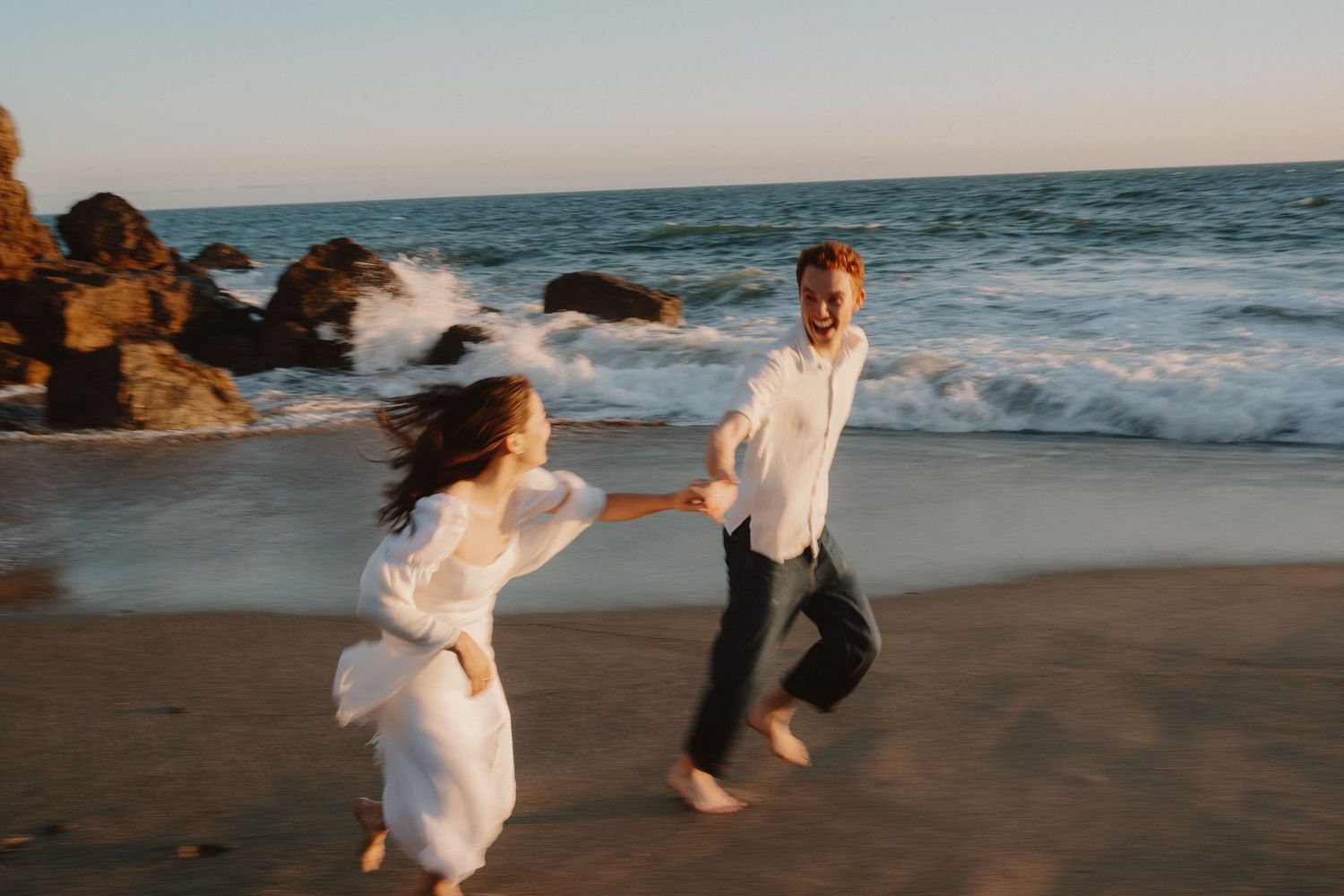 Couple playfully dancing and holding hands on a beach at golden hour with waves crashing.