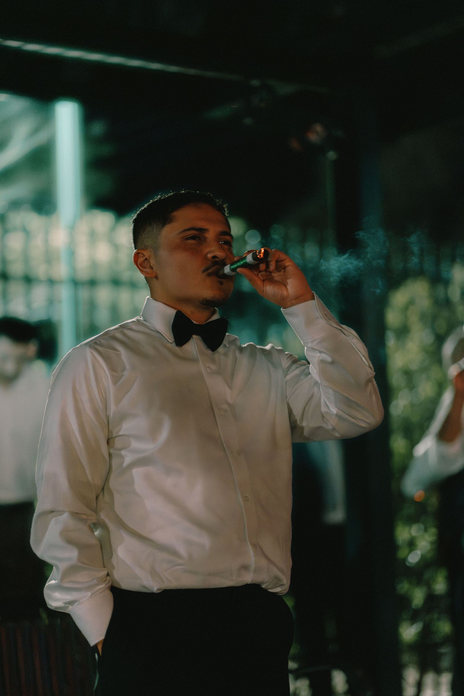 Groom in white dress shirt and bow tie smoking a cigar at evening outdoor wedding celebration.