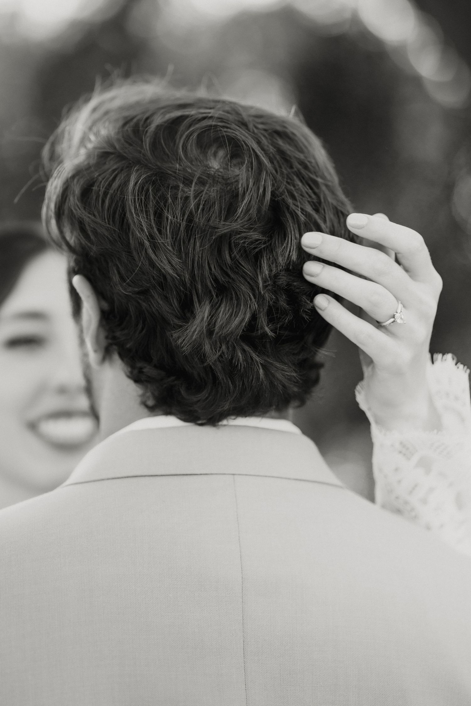 Black and white close-up of bride's hand with engagement ring running through groom's curly hair.