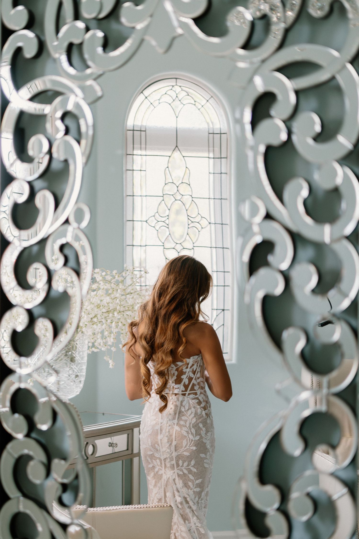 Bride in lace gown viewed through ornate mirror frame standing before a stained glass window.
