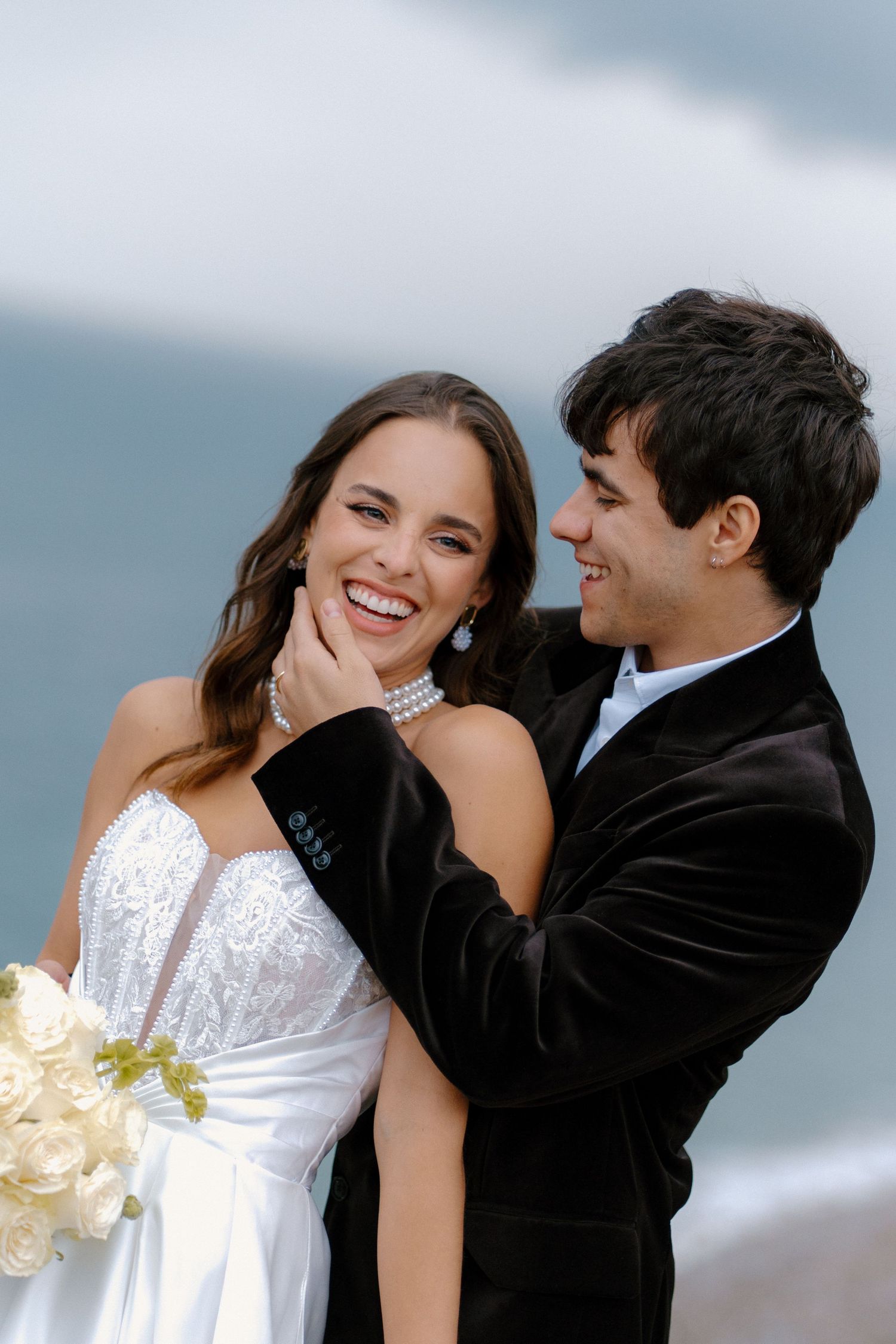Smiling groom tenderly cups brides face as she laughs joyfully, both in elegant wedding attire by the sea.