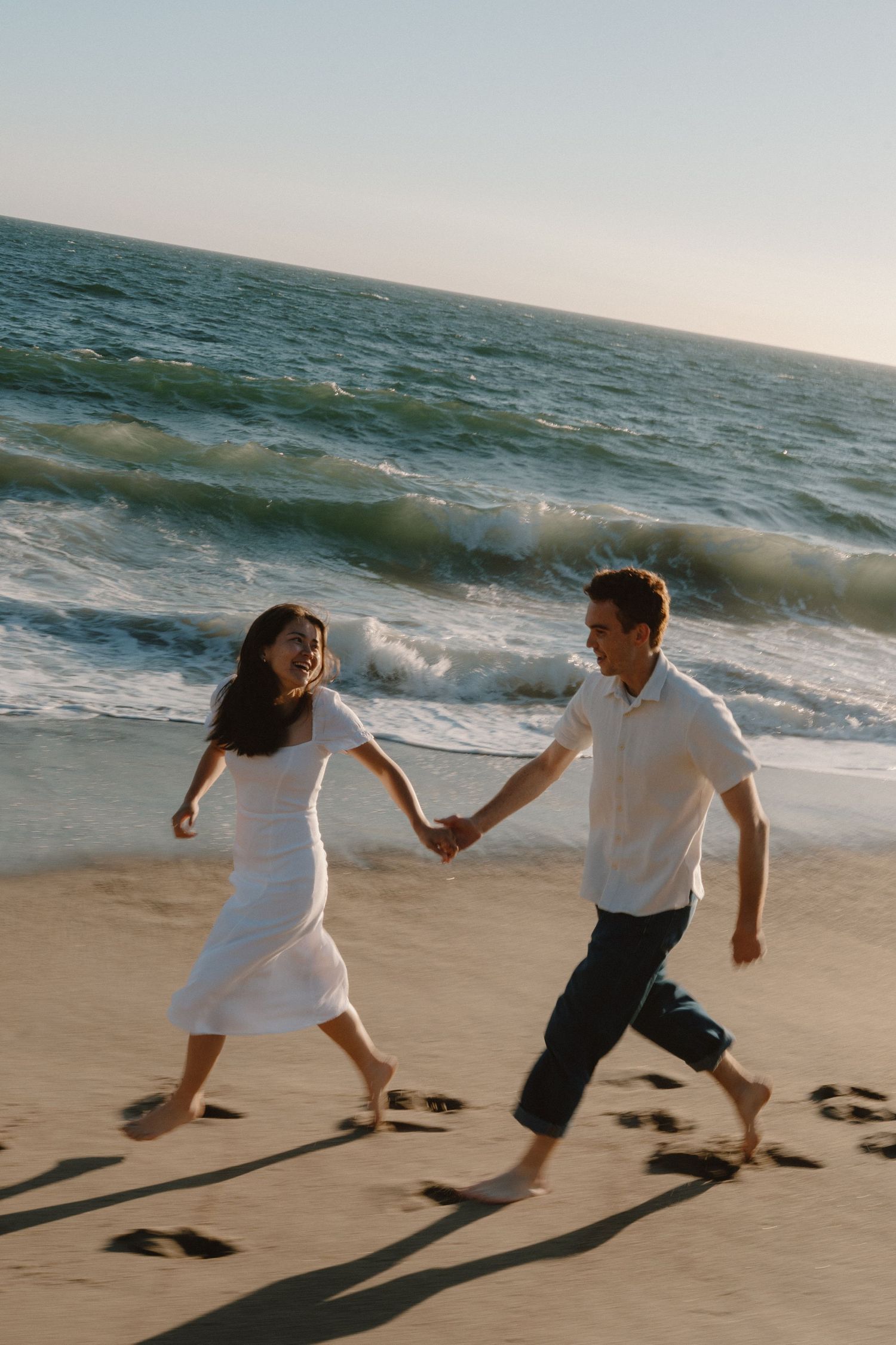 Joyful couple holding hands and running barefoot along sandy beach with ocean waves in background.