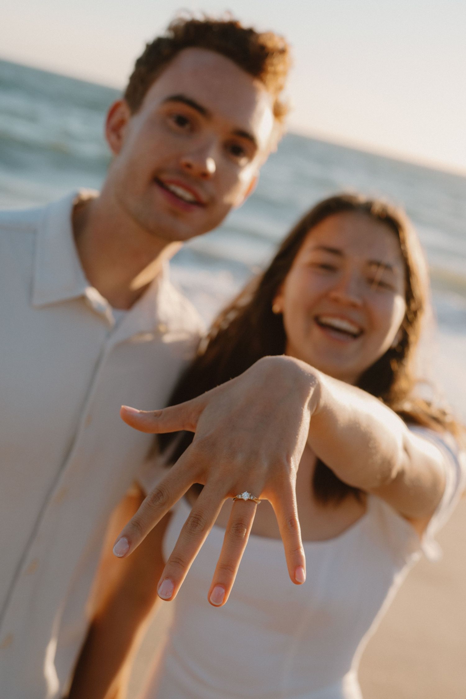 Engaged couple at beach smiling while showing off a sparkling diamond engagement ring to camera.