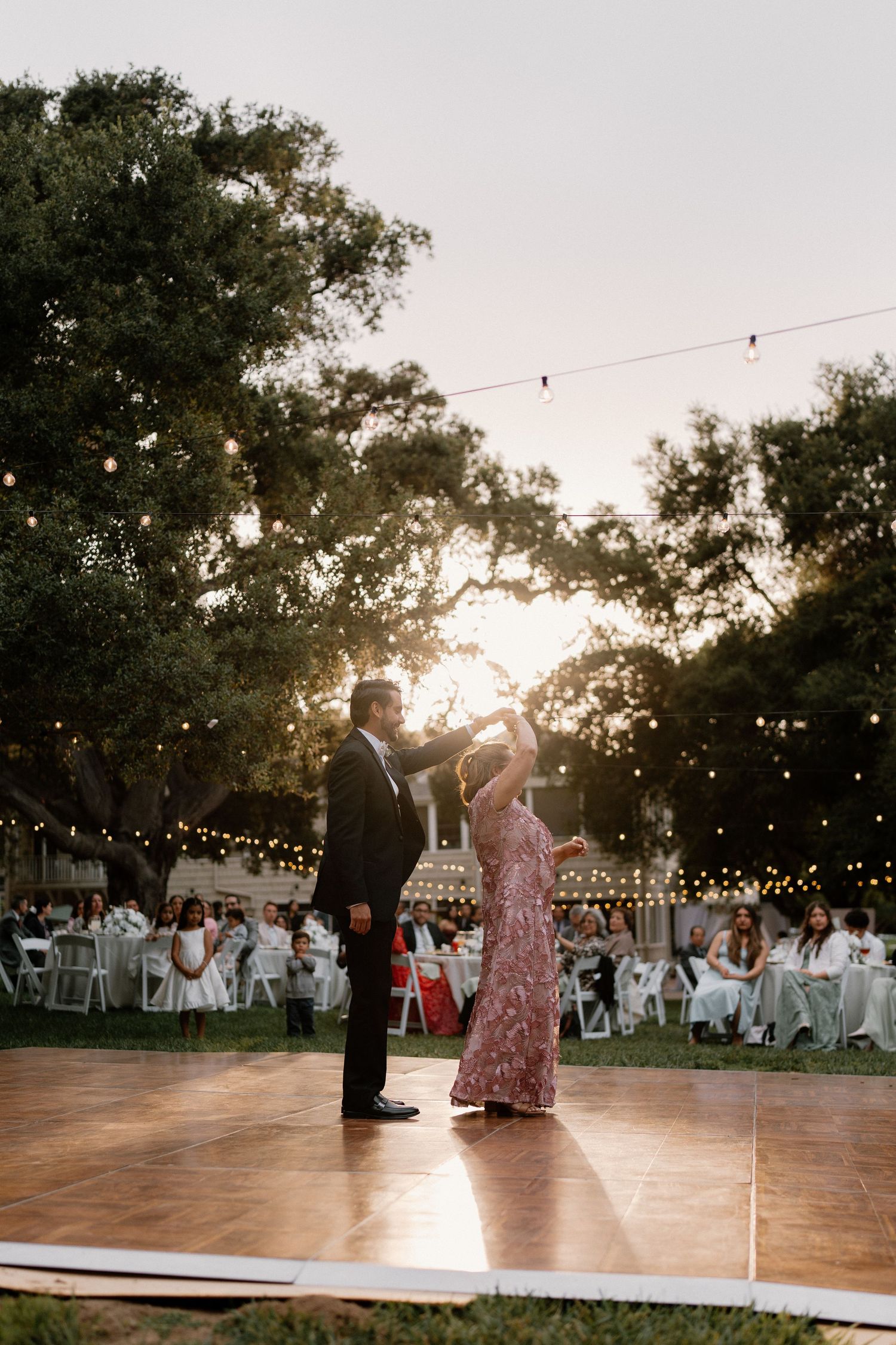 Couple shares romantic first dance on outdoor wooden floor at sunset with string lights and guests seated.