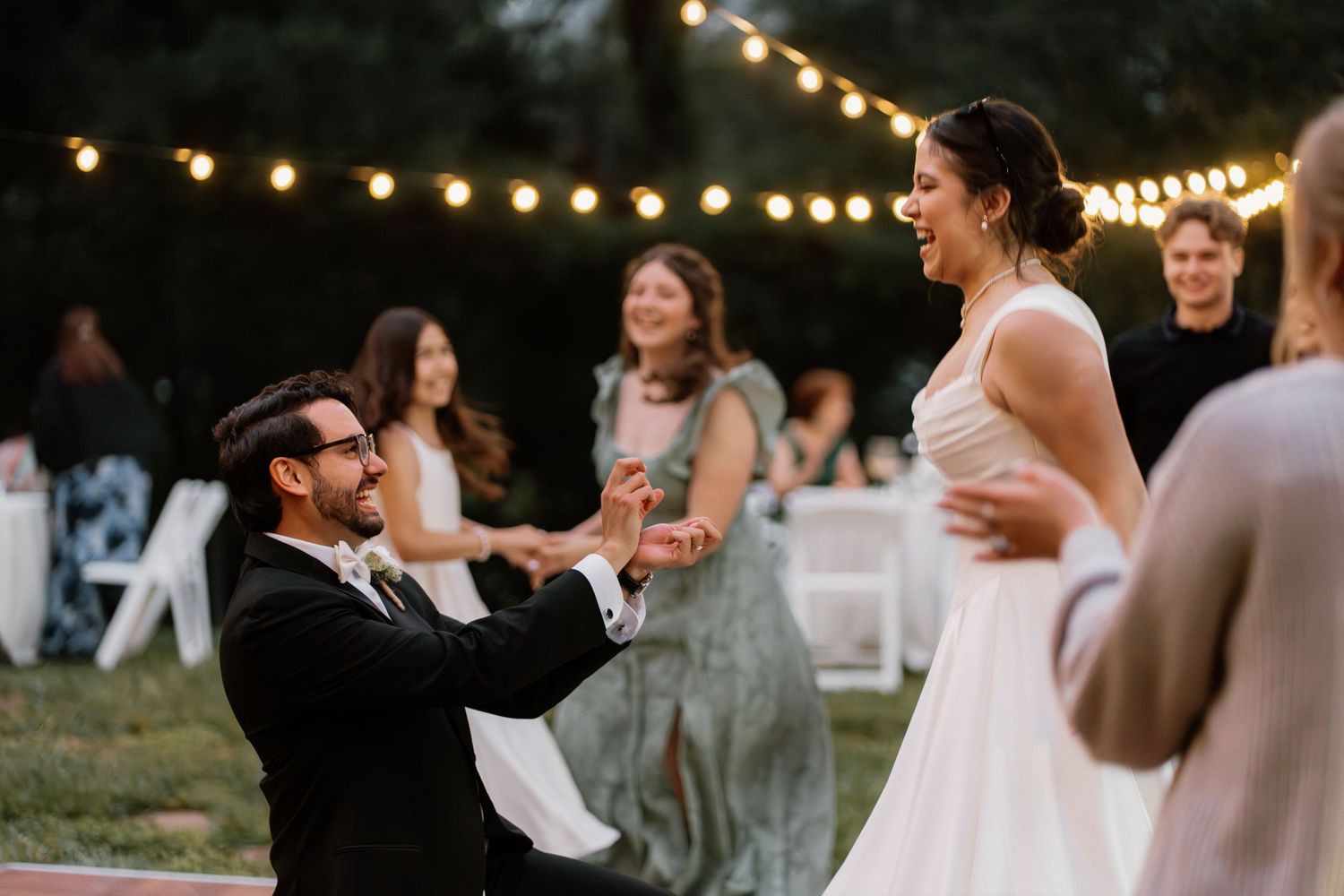 Groom kneeling before bride during outdoor wedding reception dance under string lights at dusk.