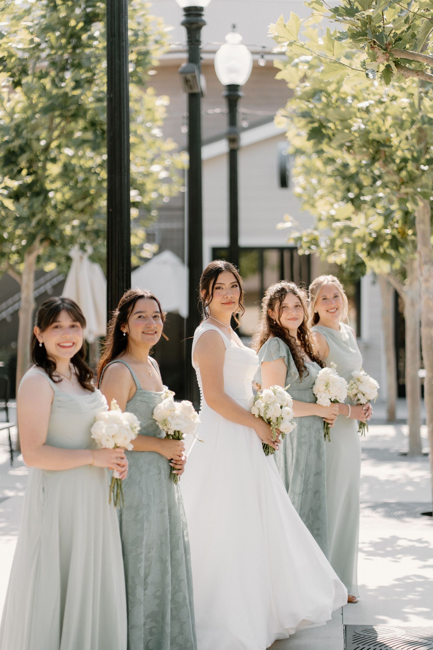 Bride in white gown poses with four bridesmaids in sage green dresses holding white bouquets outdoors.