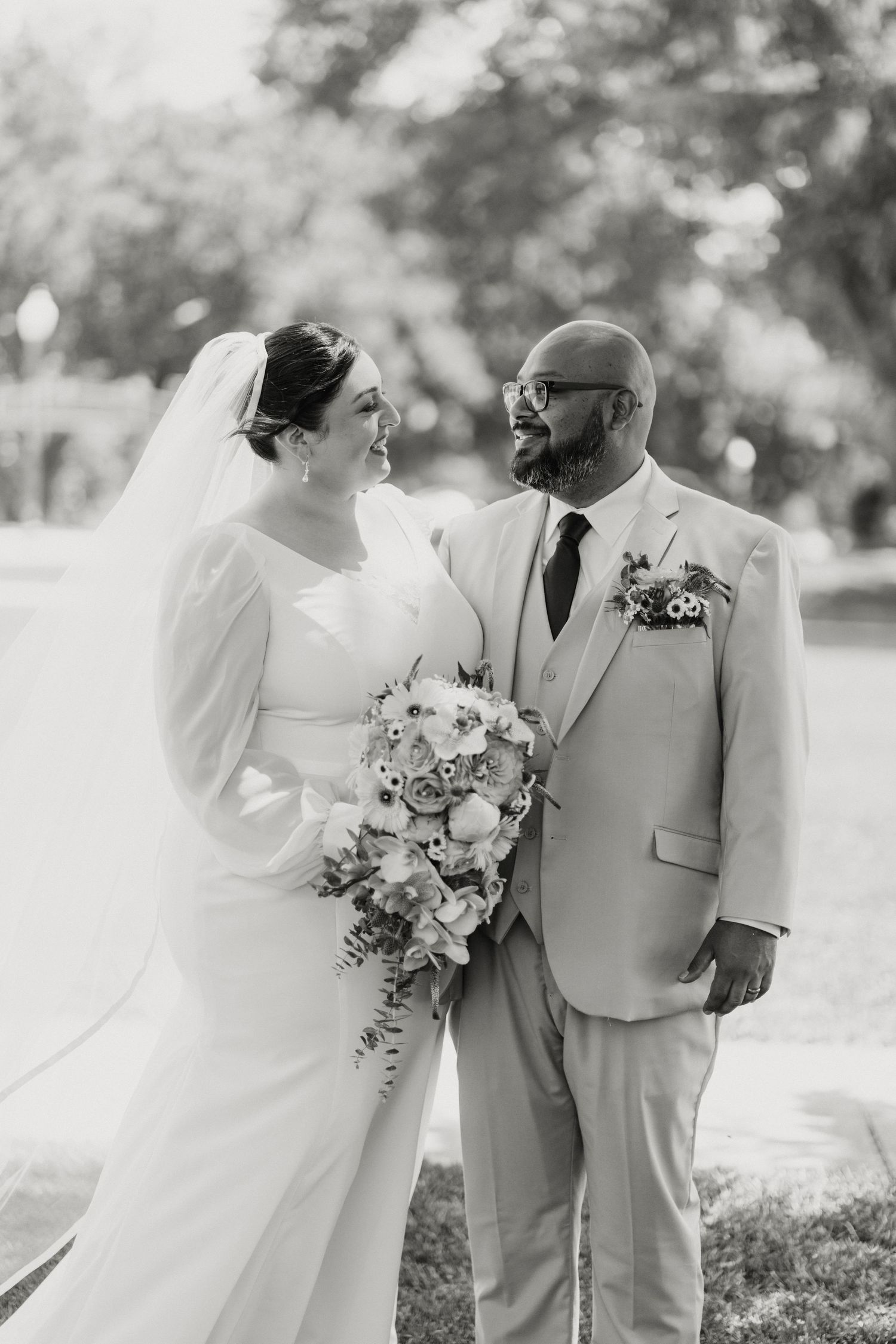 Black and white photo of bride and groom smiling at each other, bride holding colorful bouquet.
