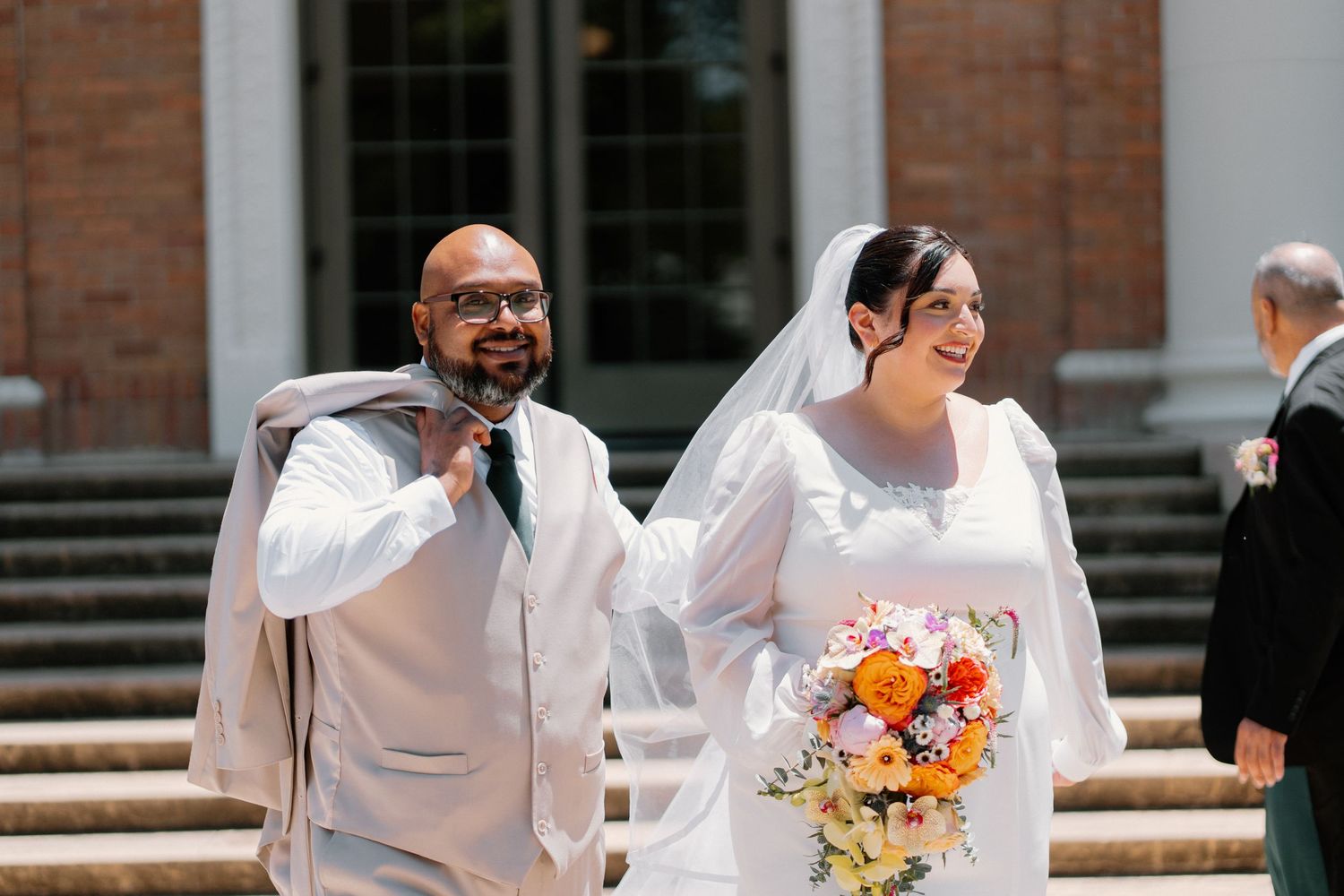 Smiling bride in white gown holding colorful bouquet walks with groom in gray suit outside brick building.
