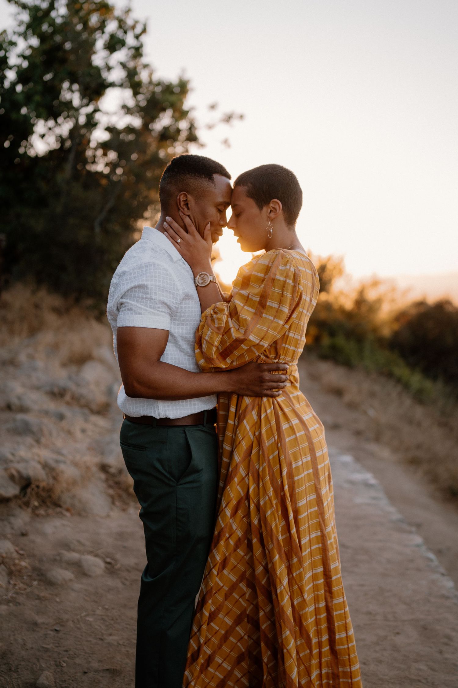 Couple sharing a romantic moment at sunset on a scenic dirt path, woman in yellow plaid dress, man in white shirt.