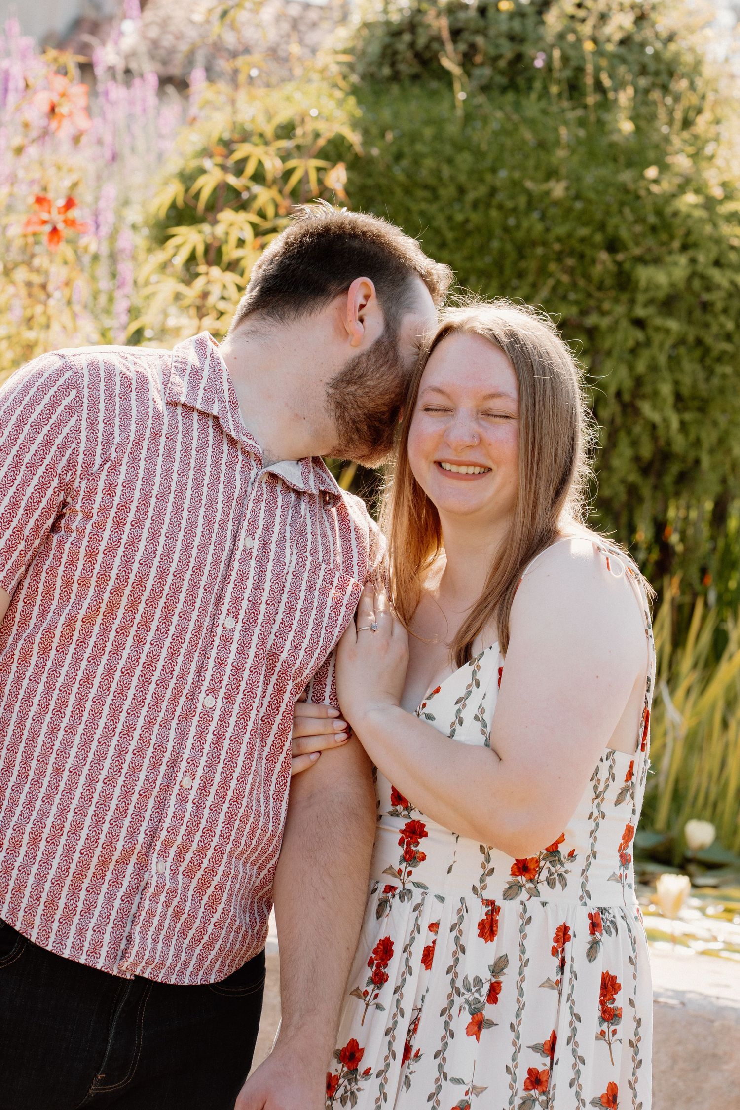 Couple sharing a tender moment outdoors, man kissing woman's cheek as she smiles in a floral dress.