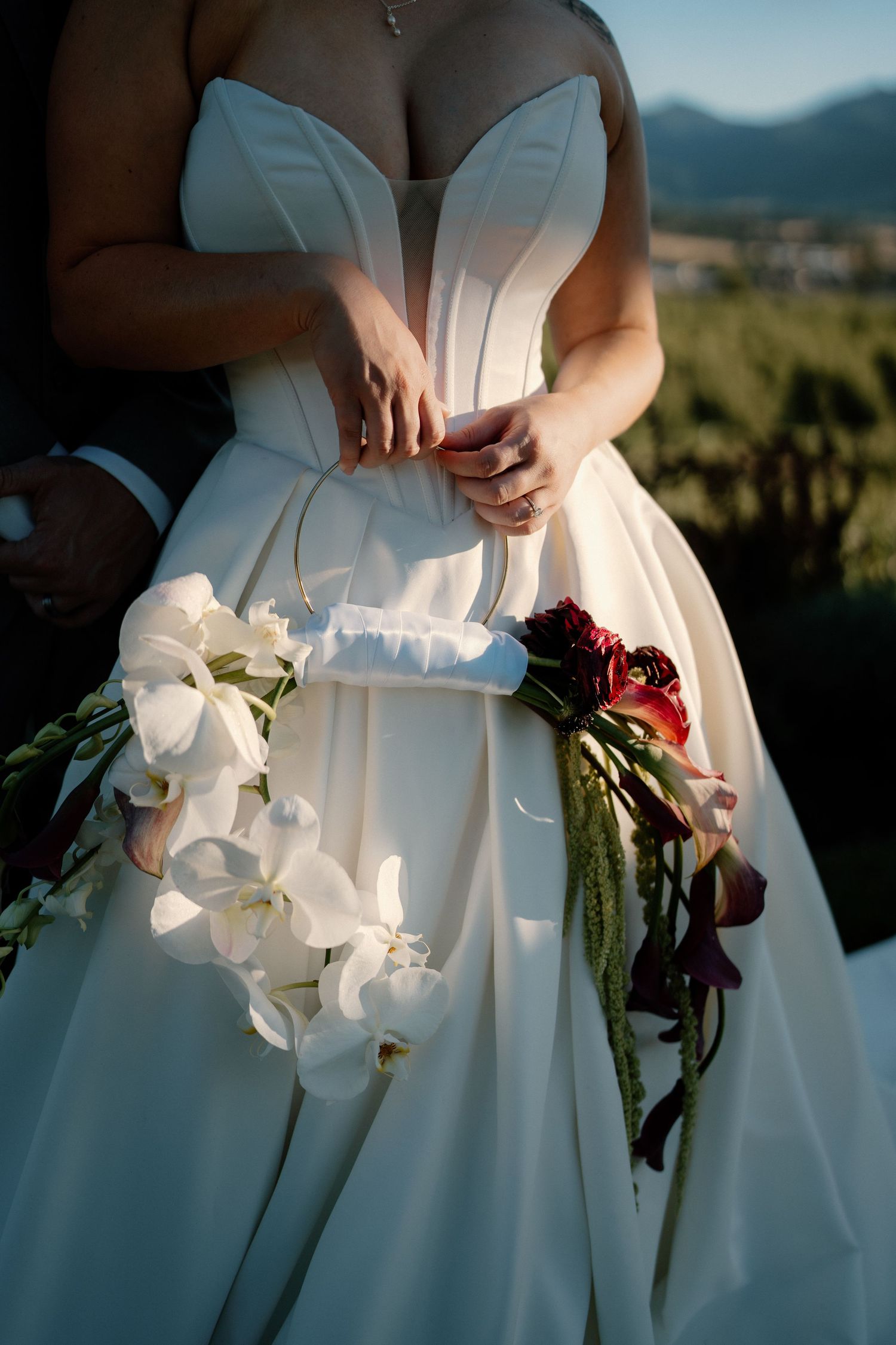 Bride in white strapless gown holding two bouquets, white orchids and red florals, with mountain views behind.