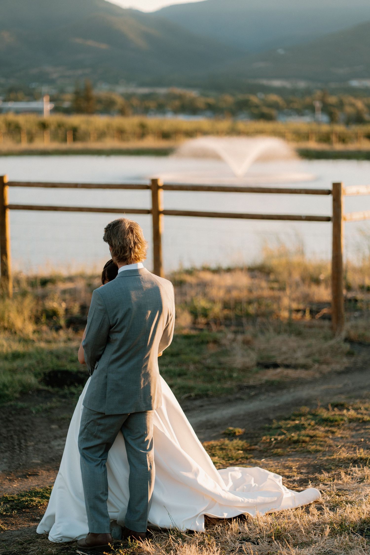 Bride and groom walk hand in hand toward a wooden fence overlooking a lake fountain at golden hour.