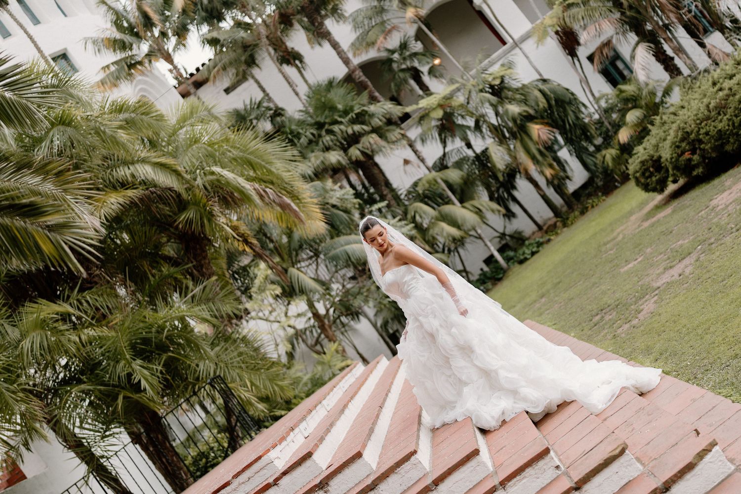 Bride in full ballgown and veil posing on steps surrounded by tropical palm trees.