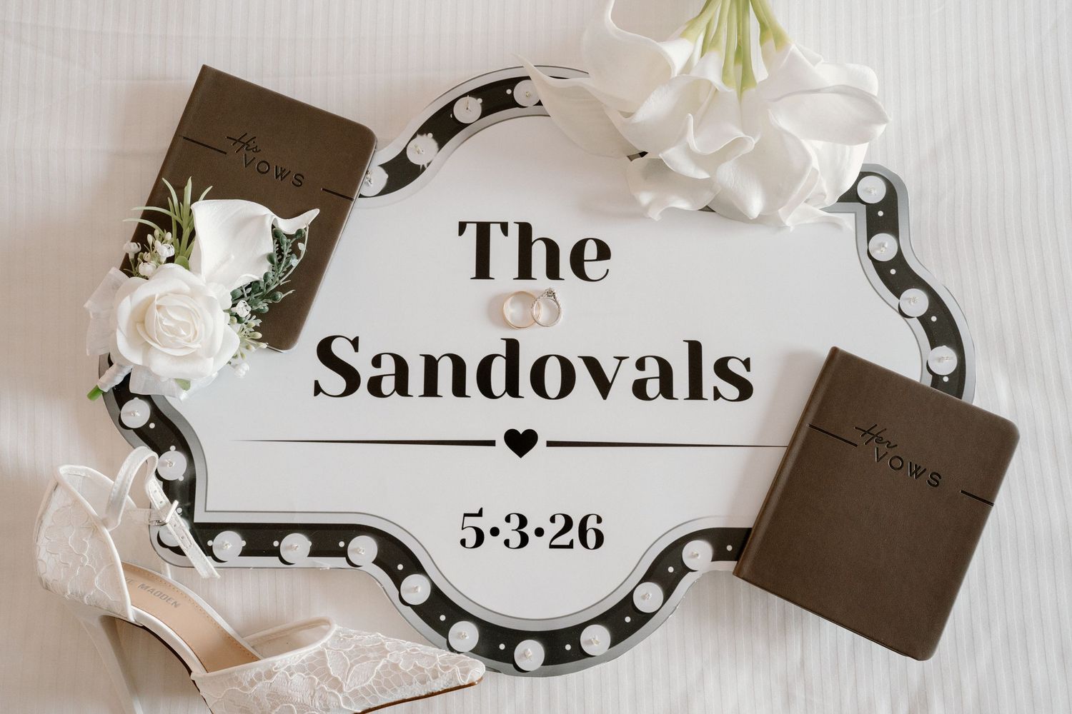 The Sandovals 5-3-26 wedding flat lay with rings, heels, vow books, and white florals.