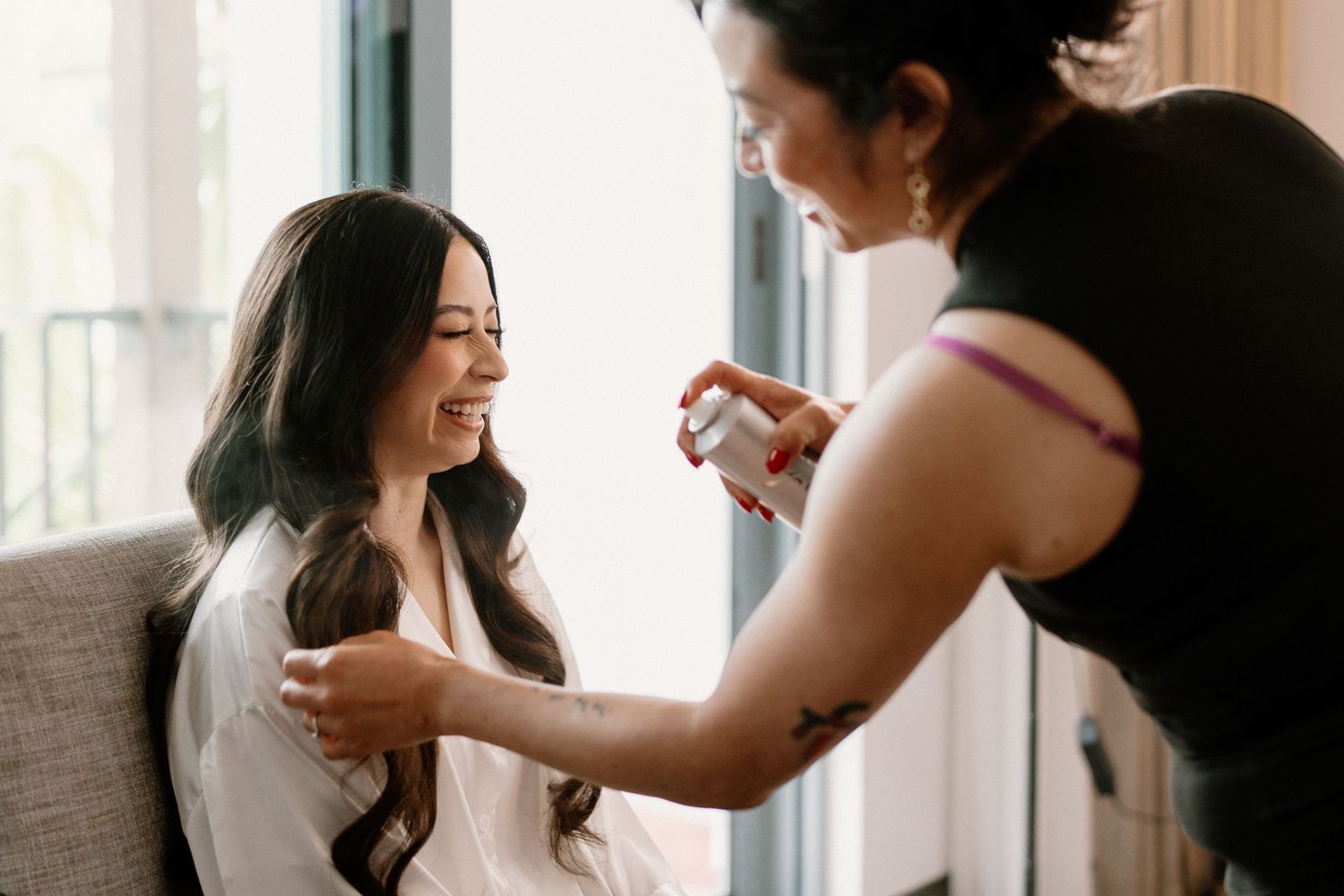 Bride smiling joyfully as hairstylist applies hairspray during wedding preparation getting ready moment.