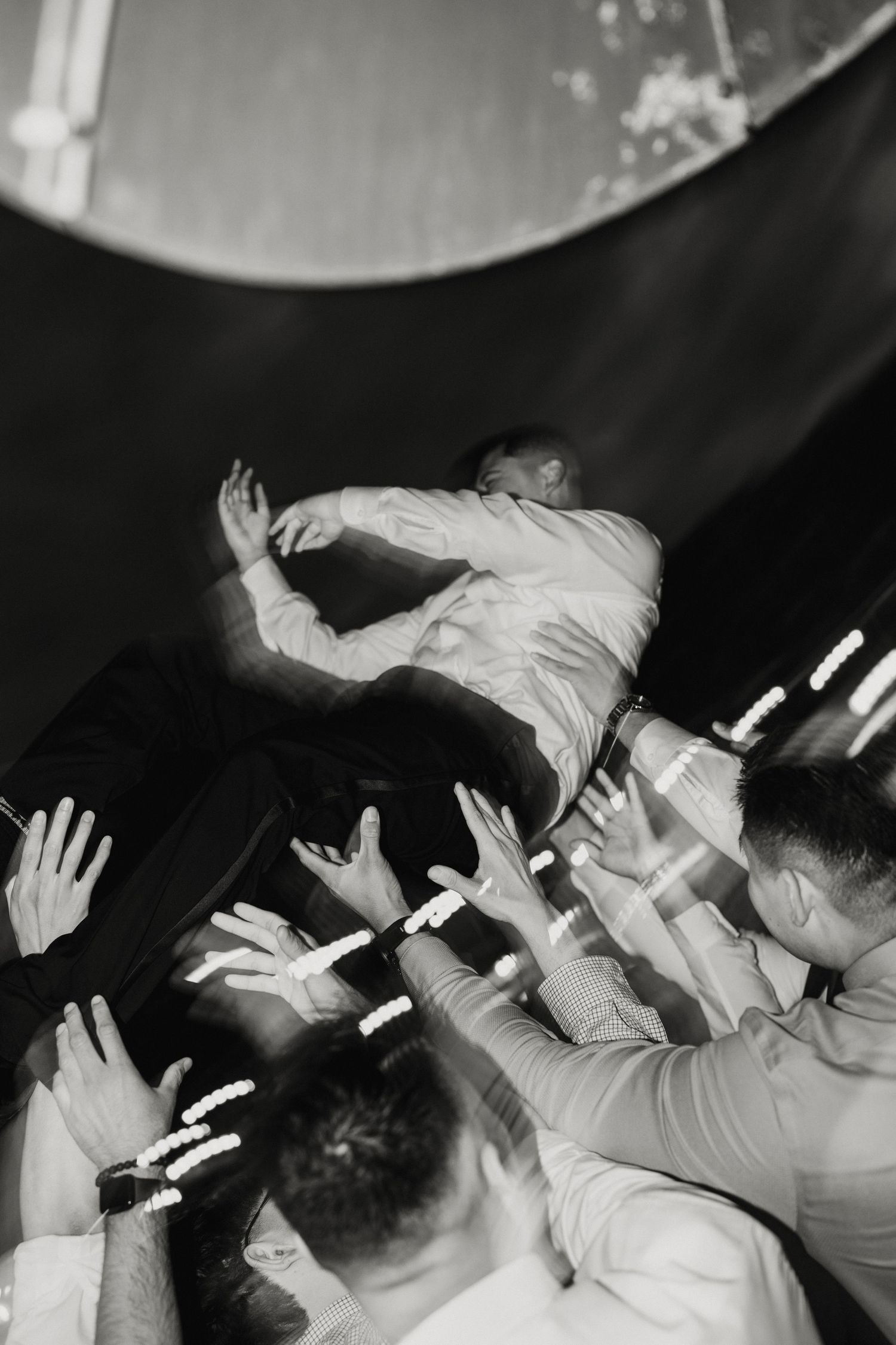 Black and white photo of a groom being lifted by wedding guests in an energetic crowd surf celebration.
