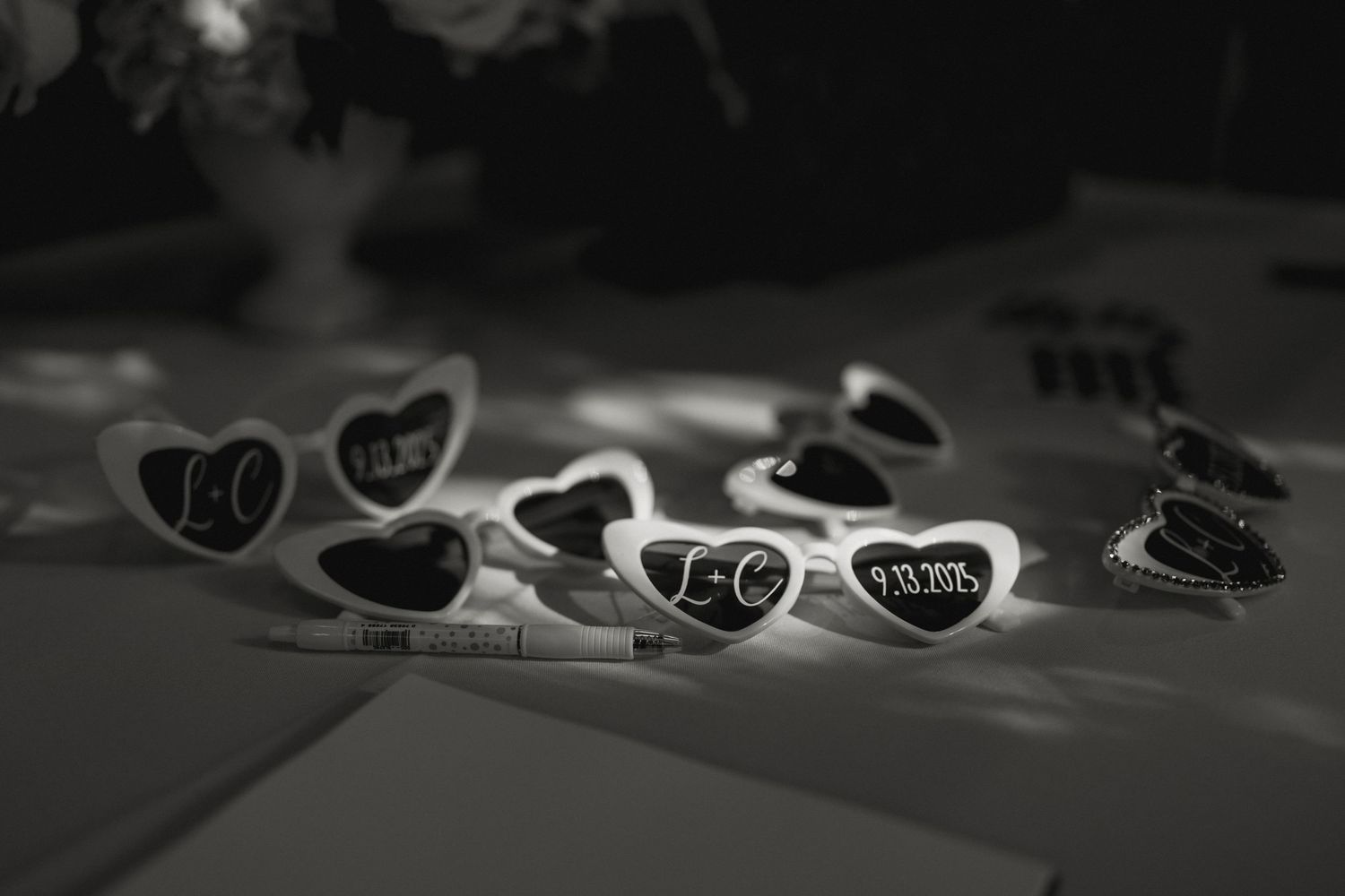 Black and white photo of heart-shaped sunglasses with initials as fun wedding party favors on a table.