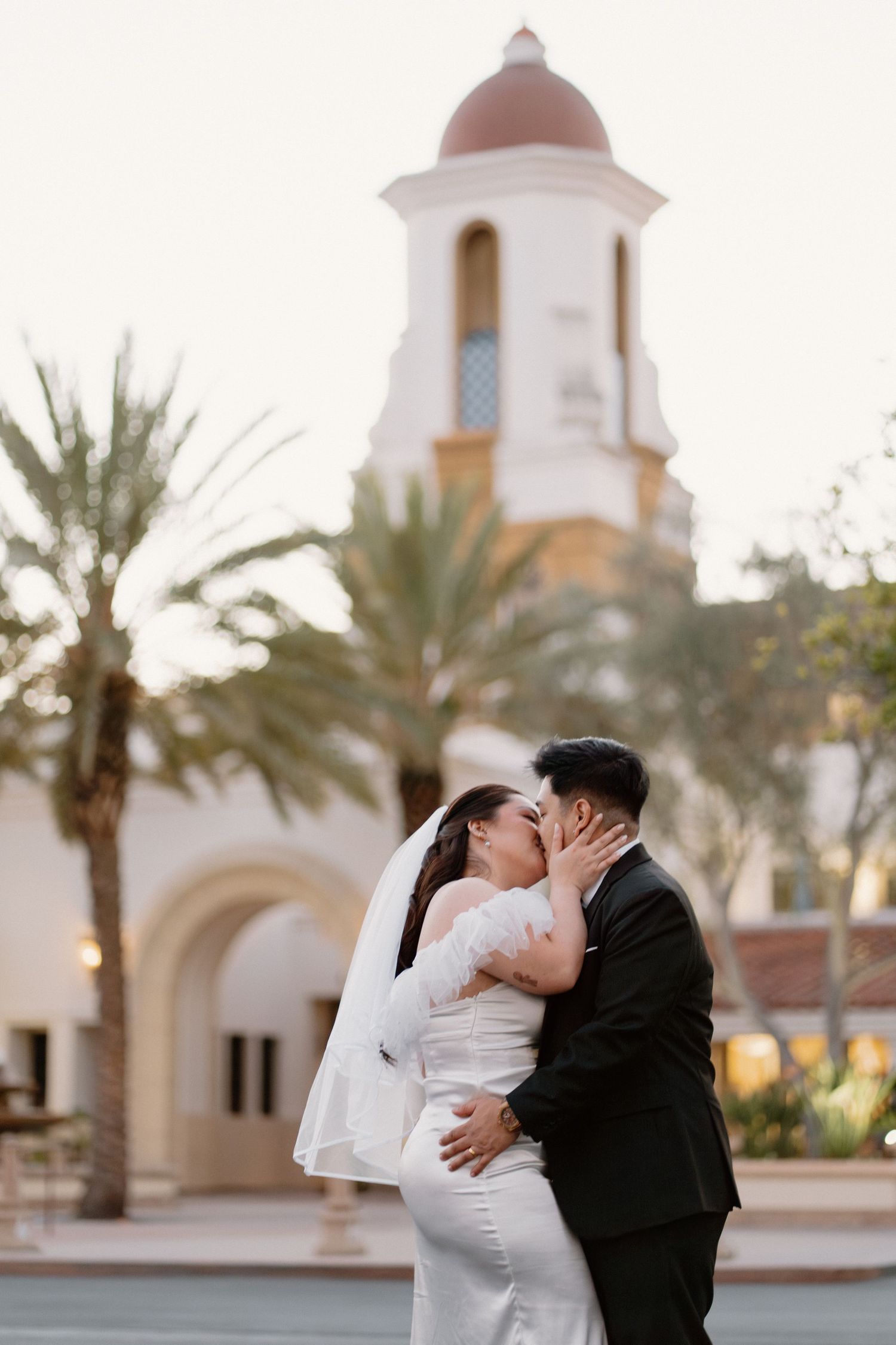Asian bride and groom sharing a kiss in front of a white church with palm trees.