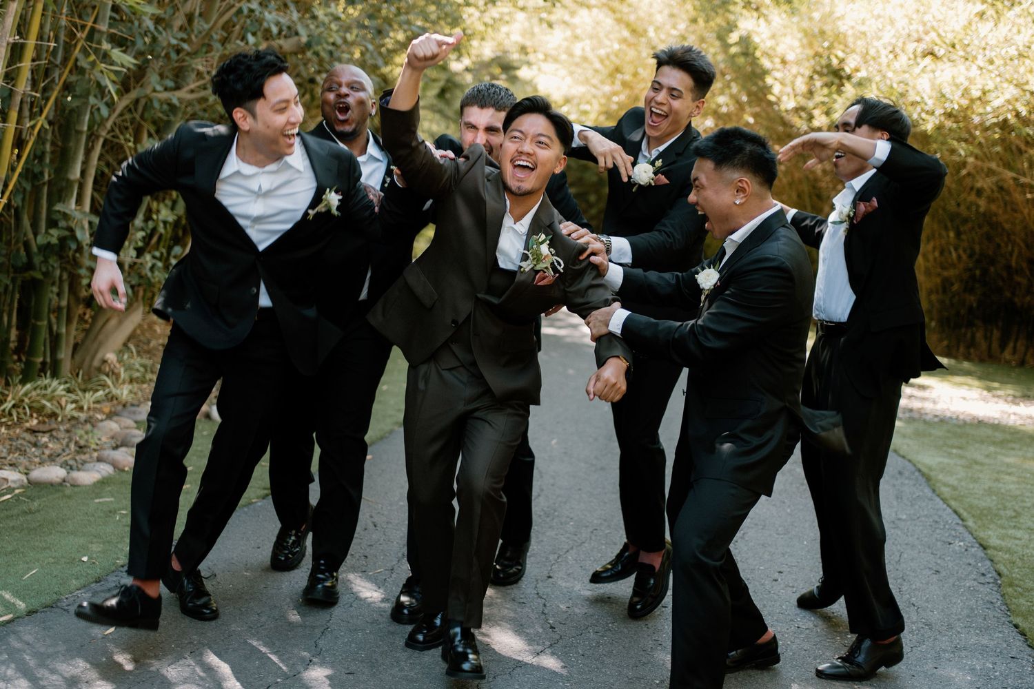 Excited groom celebrated by cheering groomsmen in black suits along bamboo-lined pathway.