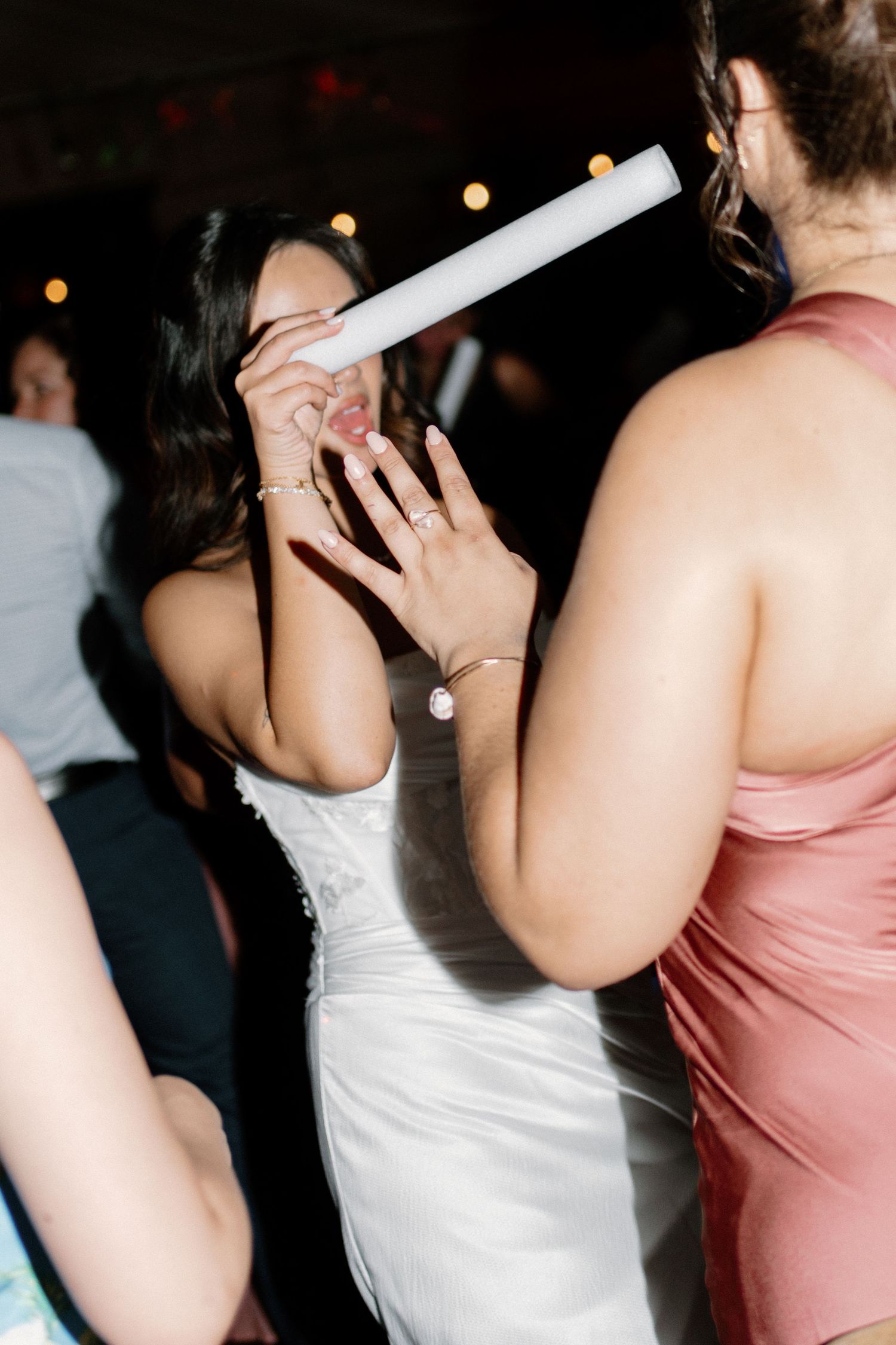 Bride laughs while dancing with guests at lively wedding reception under warm string lights.