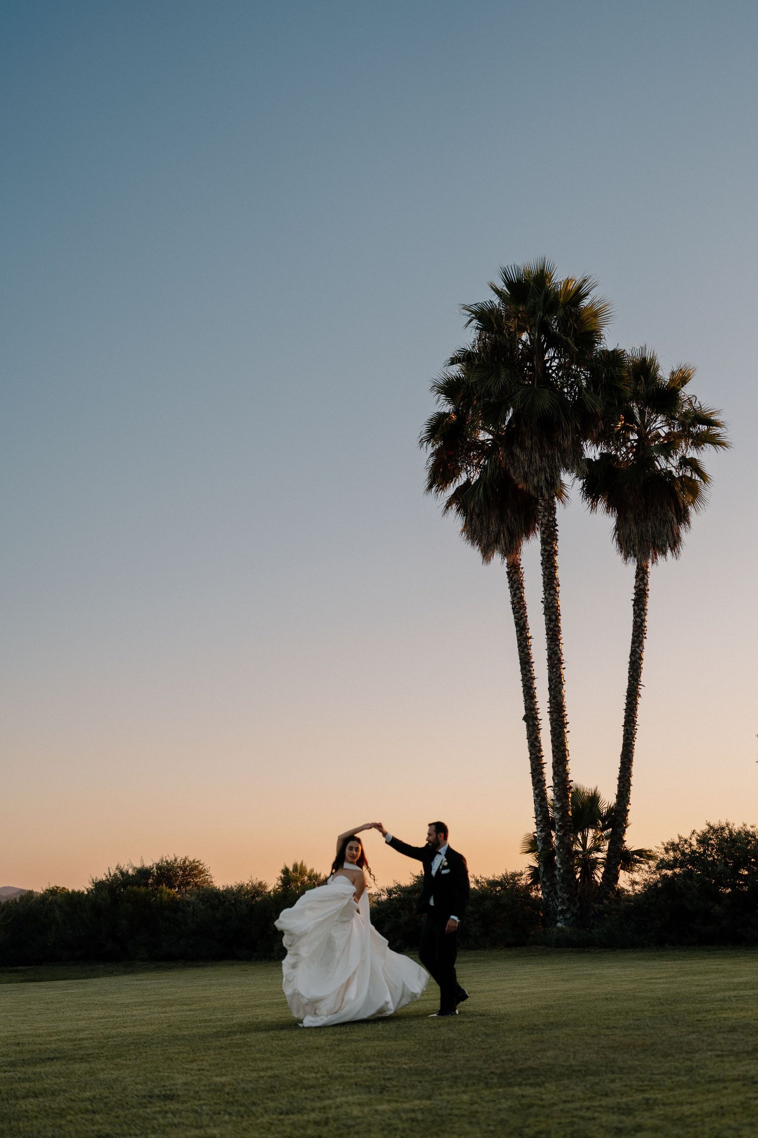 A bride and groom dance at sunset beside tall palm trees on a lush green lawn.