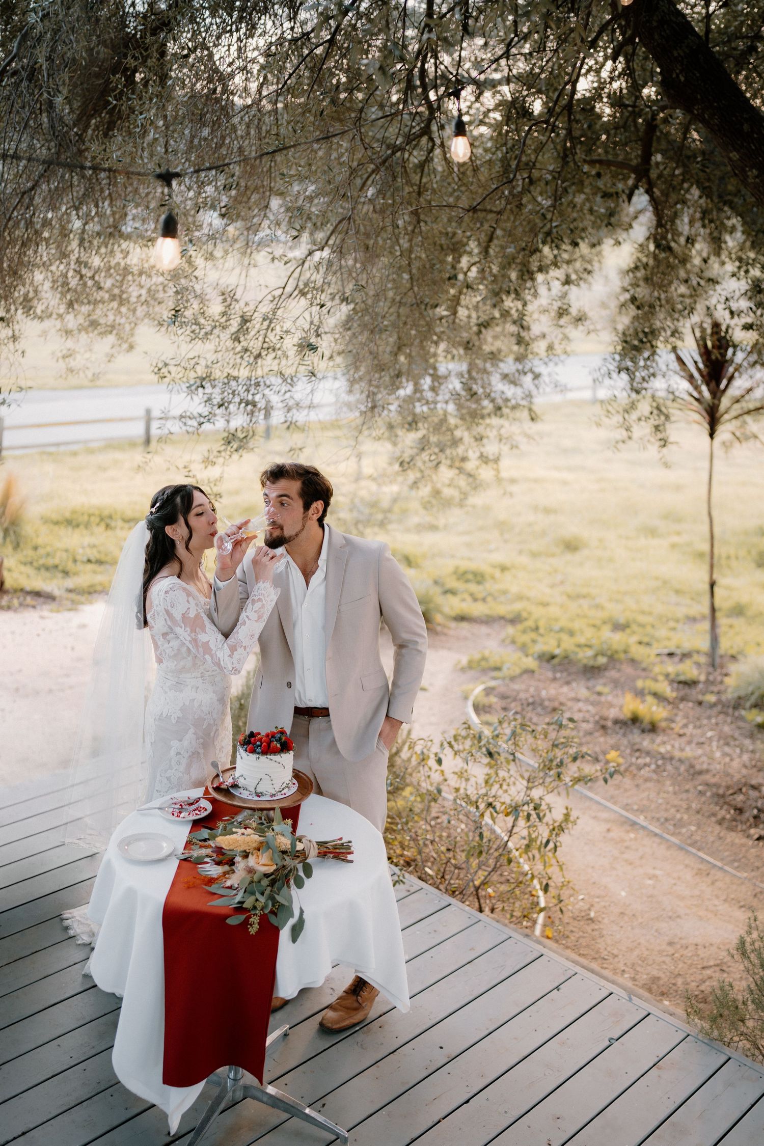 Bride and groom sharing wedding cake at outdoor table with red runner under string lights at dusk.