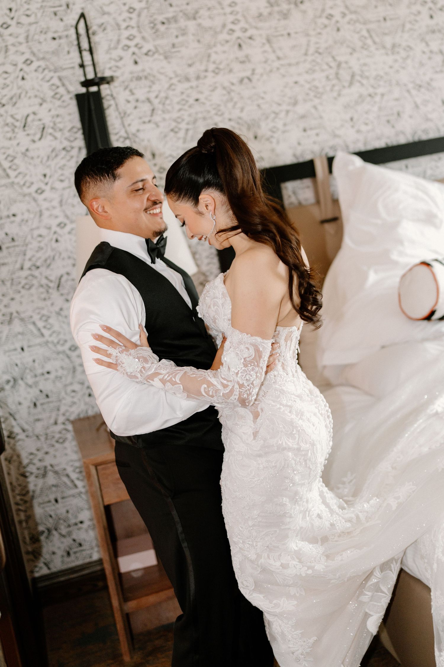 Bride in off-shoulder lace gown and groom in black vest share a romantic embrace and smile at each other.