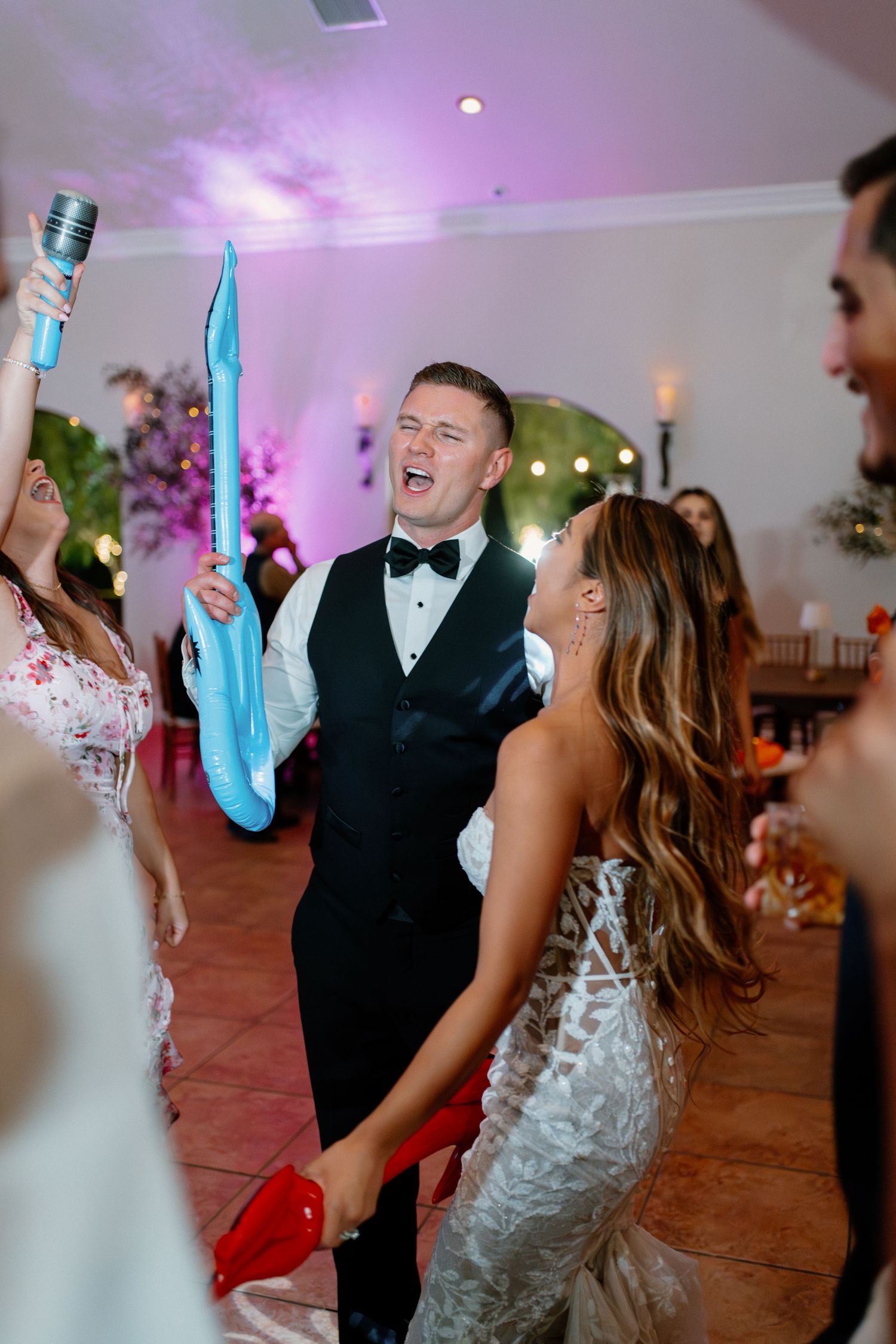 Bride and groom dancing joyfully at wedding reception holding inflatable toys under purple lights.