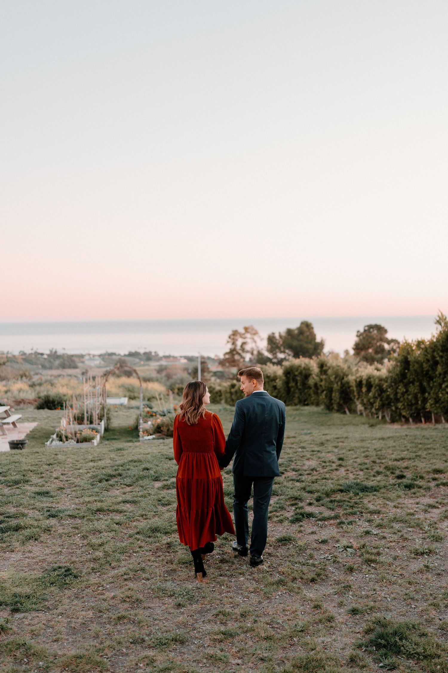 Engaged couple walks hand in hand toward ocean horizon at sunset.