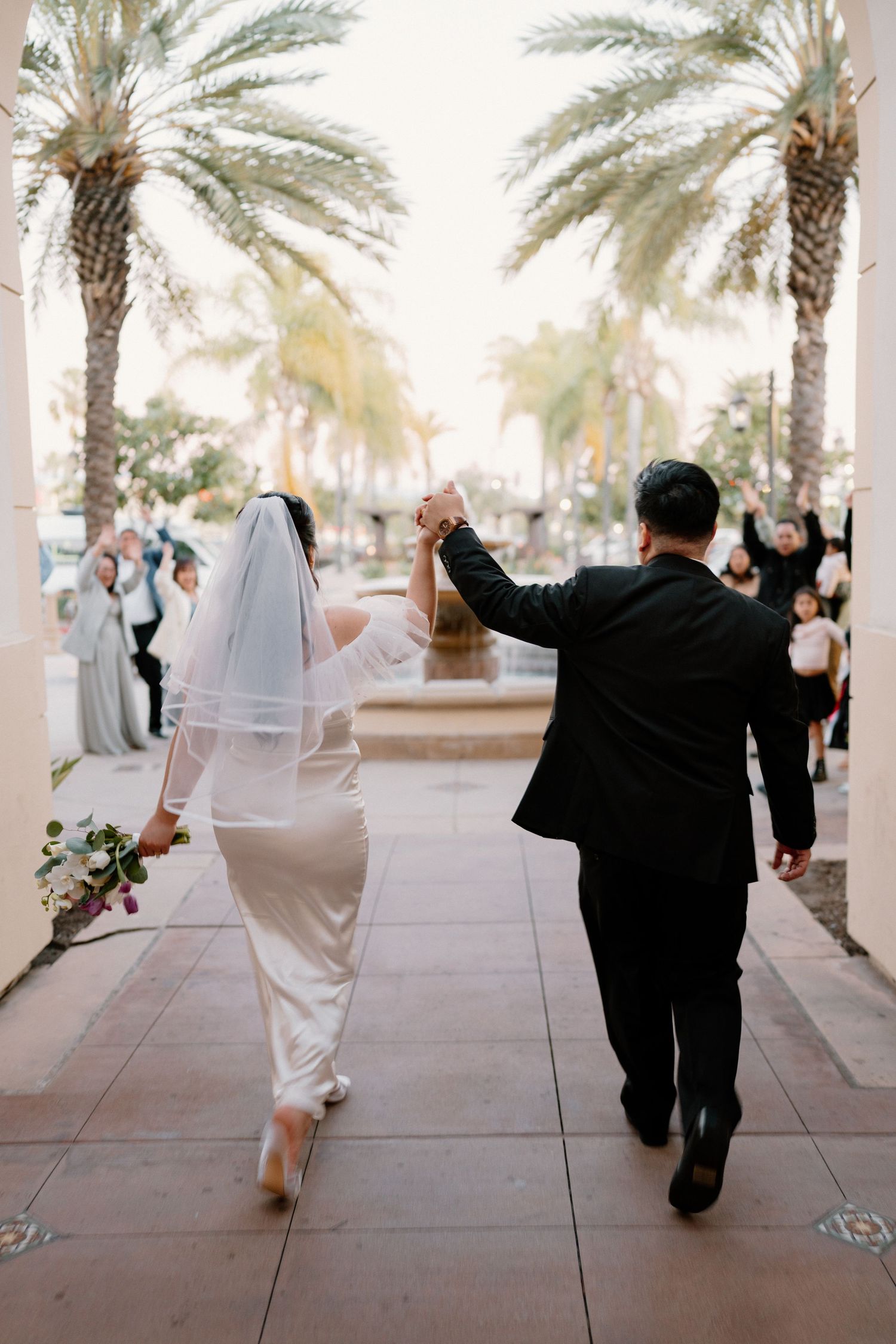 Newlyweds walk hand-in-hand under palm trees as guests celebrate their wedding exit.