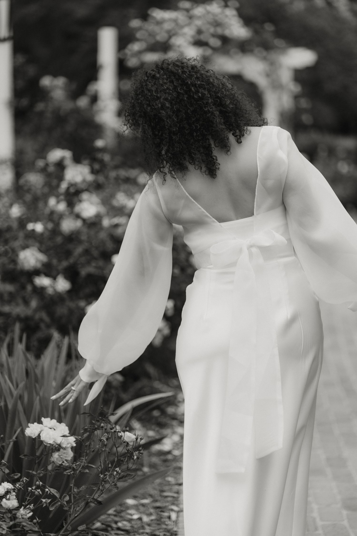 Black and white photo of bride in white dress with bow back reaching down to touch garden flowers.