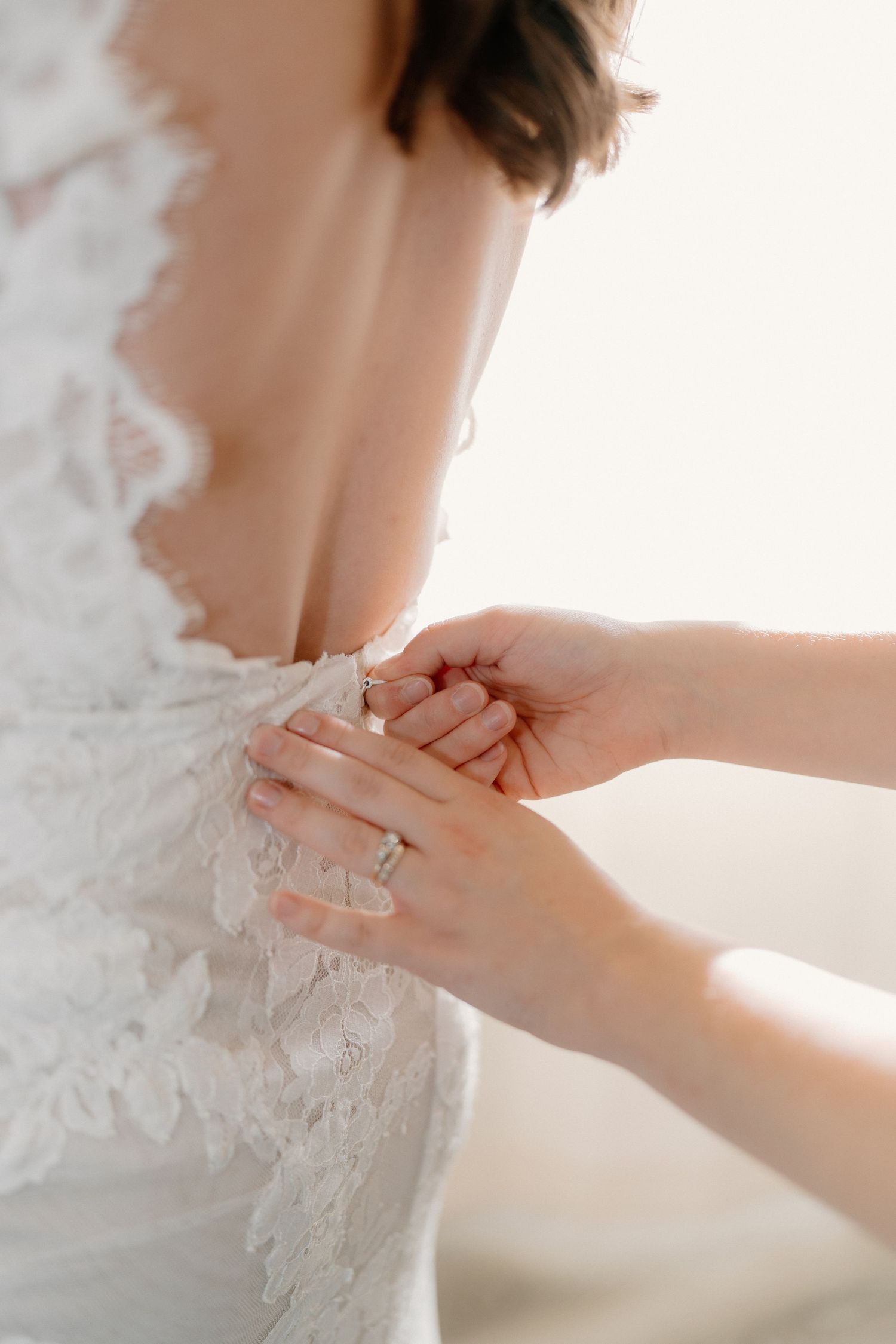 Close-up of hands fastening the back buttons of a white lace wedding dress on a bride during preparations.
