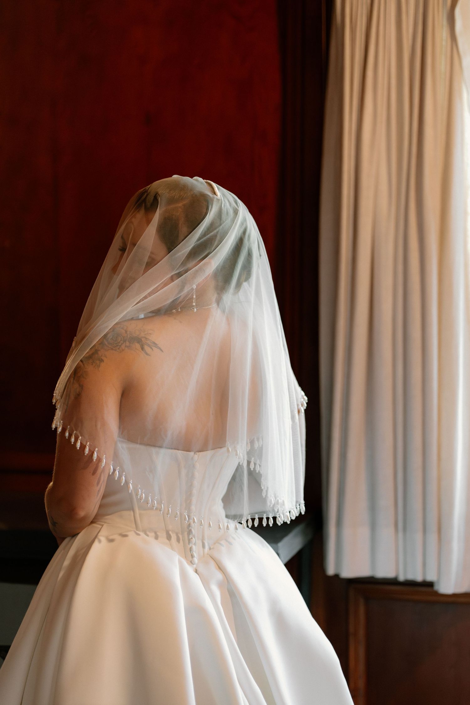 Bride in white satin wedding gown and veil seen from behind near a sunlit window.