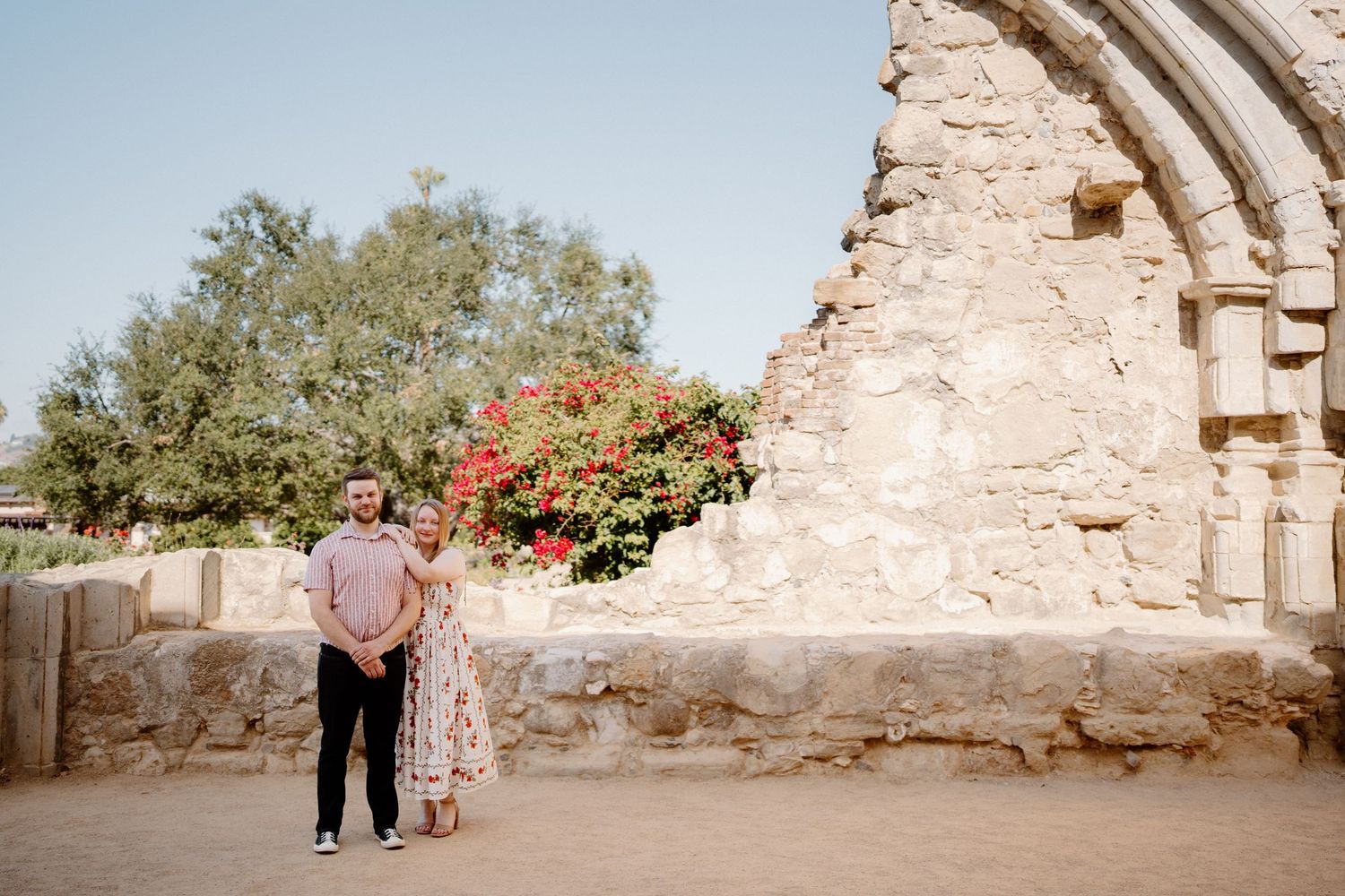 Couple standing together near ancient stone ruins with blooming red flowers and lush greenery at Mission San Juan Capistrano.
