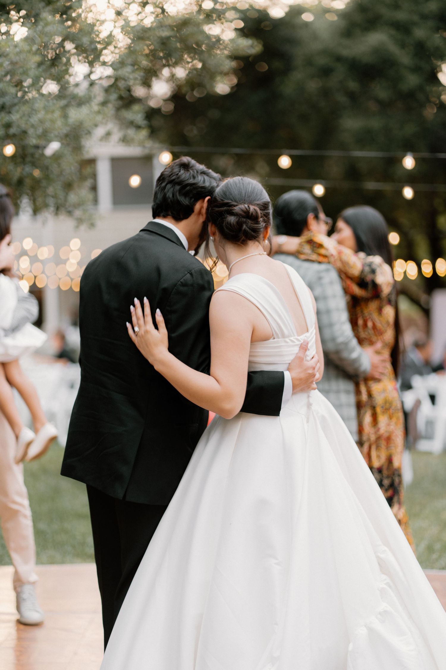 Bride and groom share first dance outdoors under string lights at evening wedding reception.