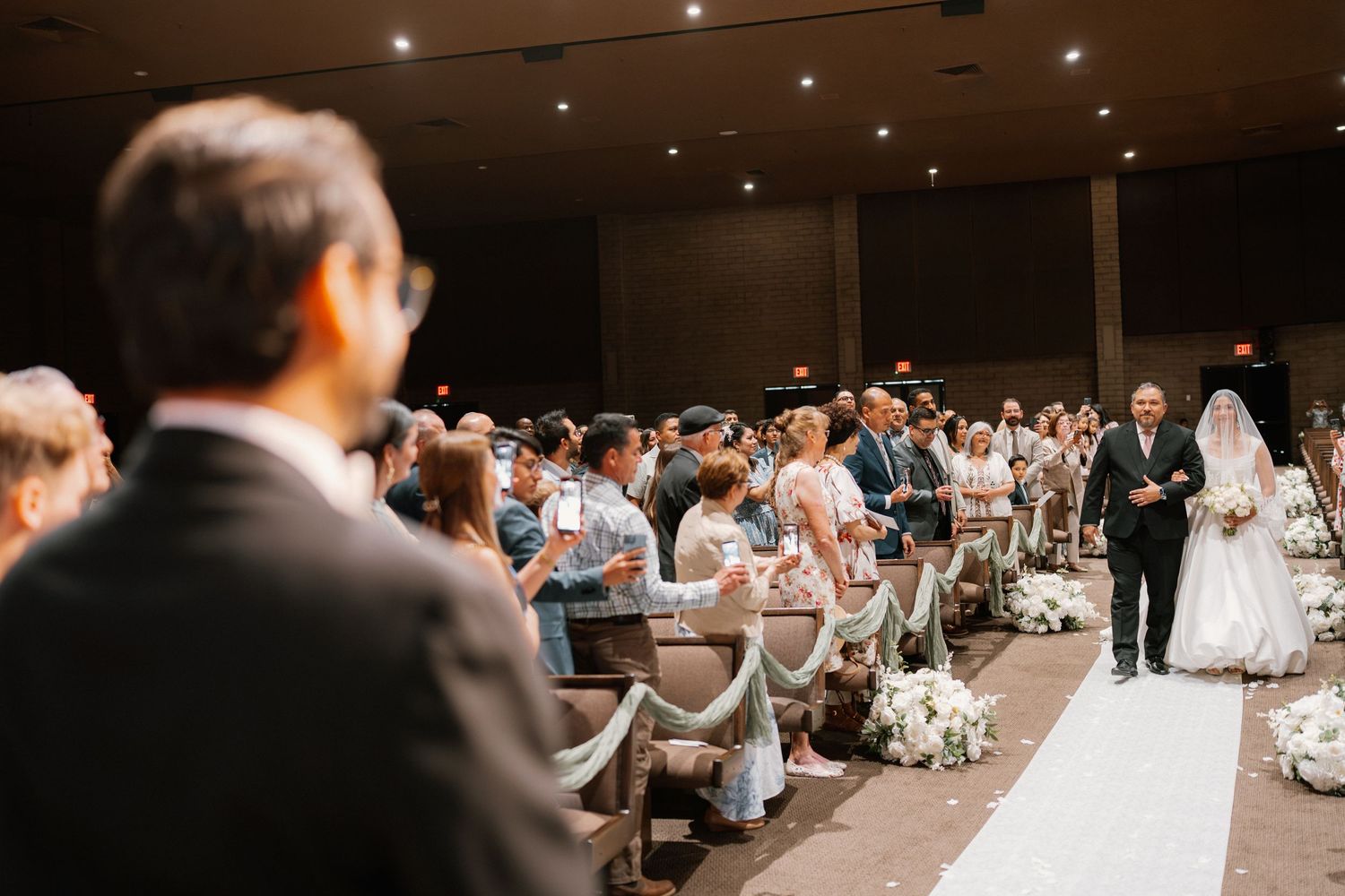 Bride walking down the aisle with escort at indoor wedding ceremony with seated guests.