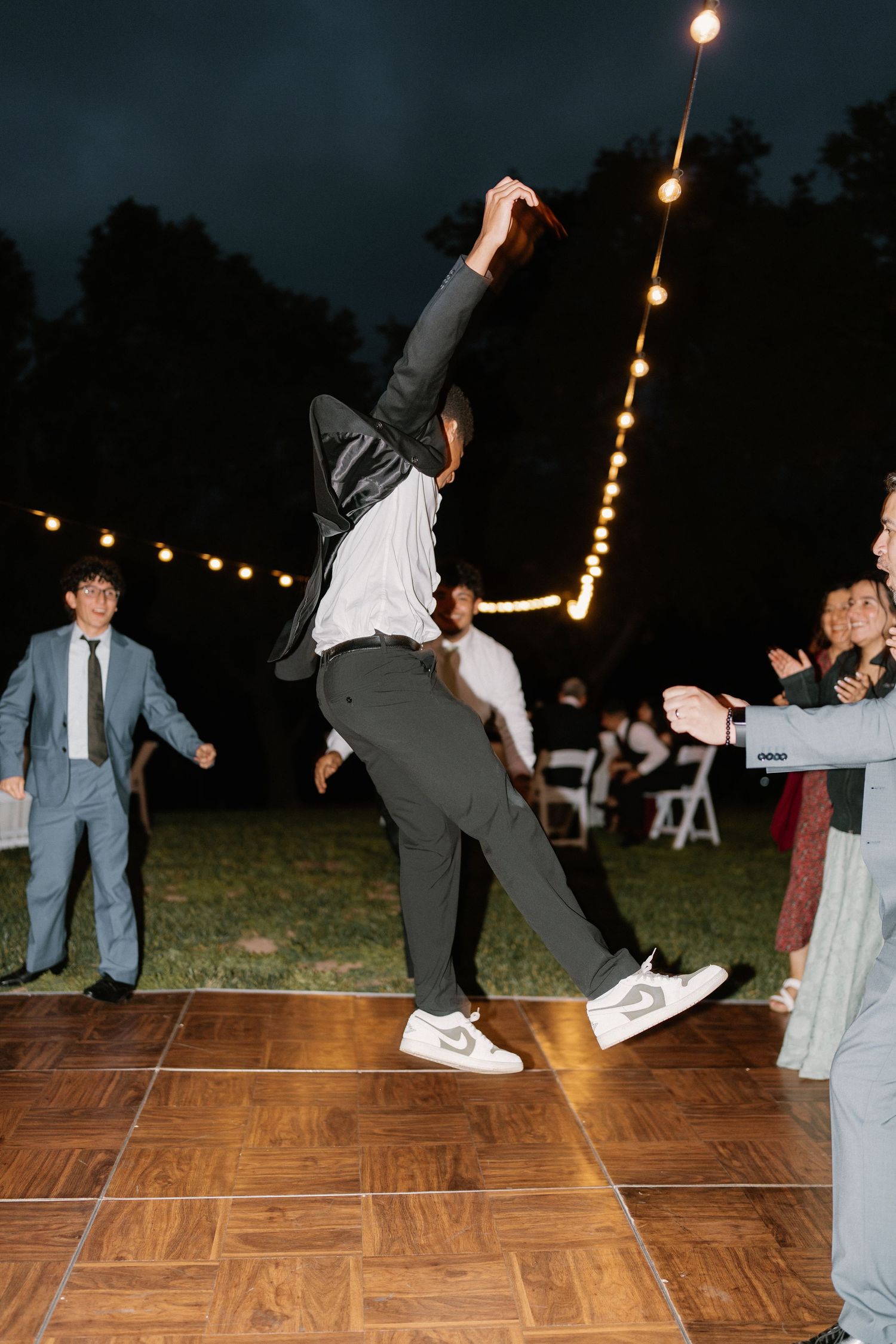 Wedding guest performs energetic jump on outdoor dance floor surrounded by cheering guests at night reception.
