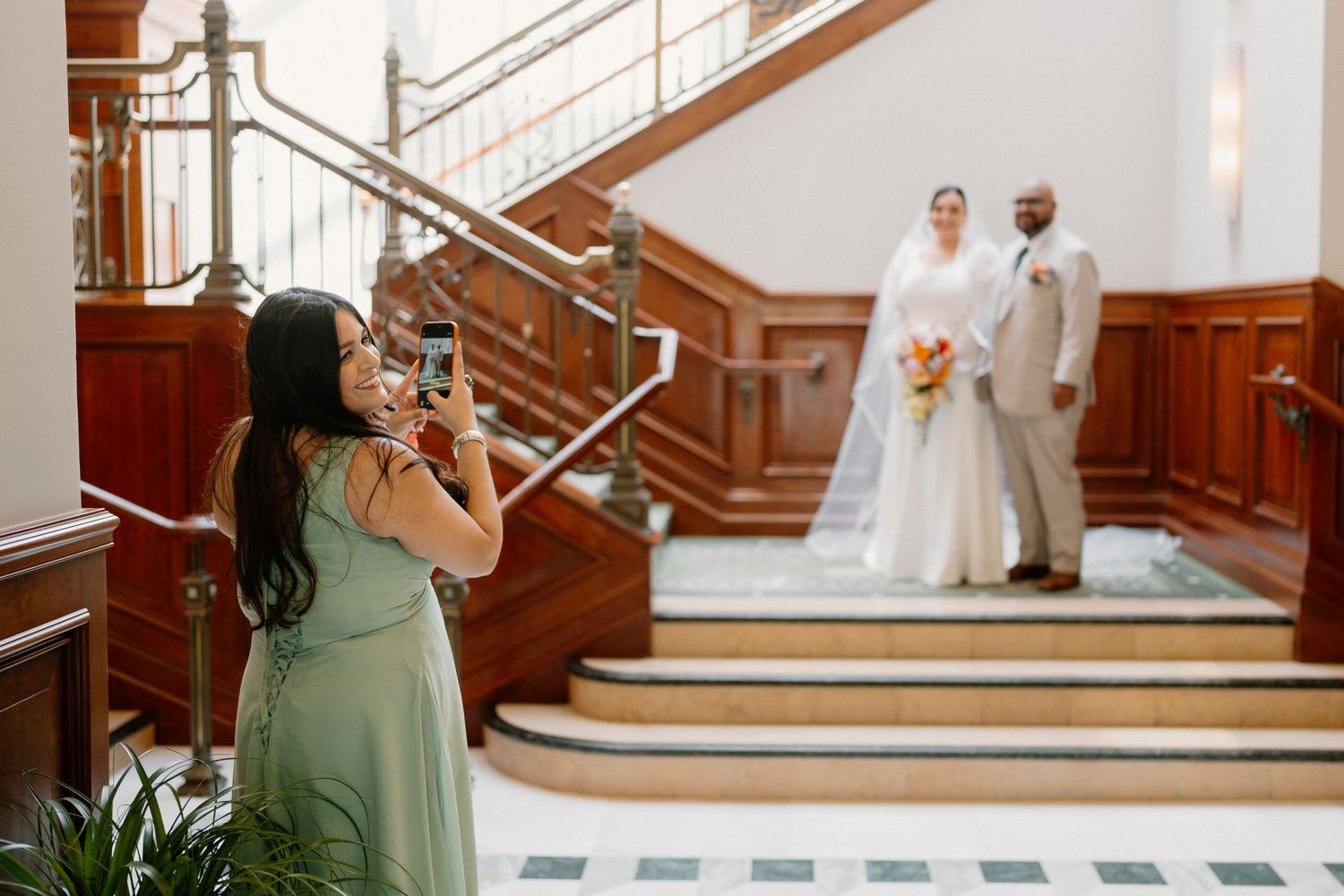Bridesmaid photographs bride and groom on grand wooden staircase at wedding venue.