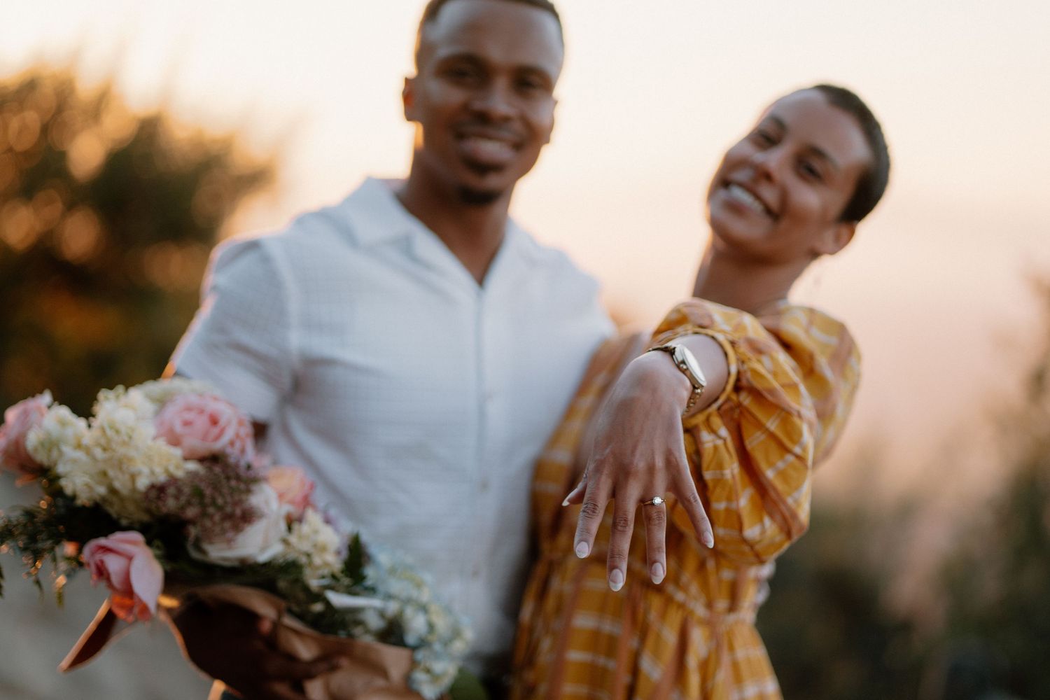 Happy couple celebrating engagement with bouquet and diamond ring at golden hour sunset.