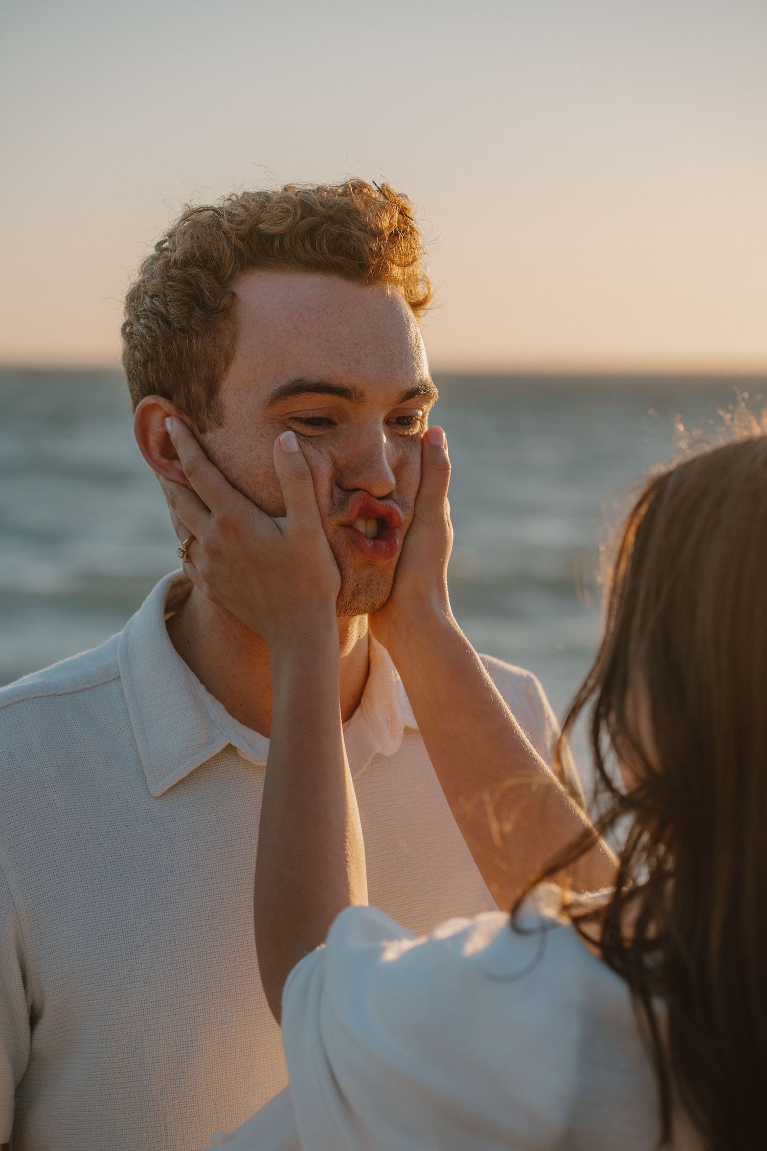 A woman tenderly touches a man's face near the ocean during golden hour sunset.