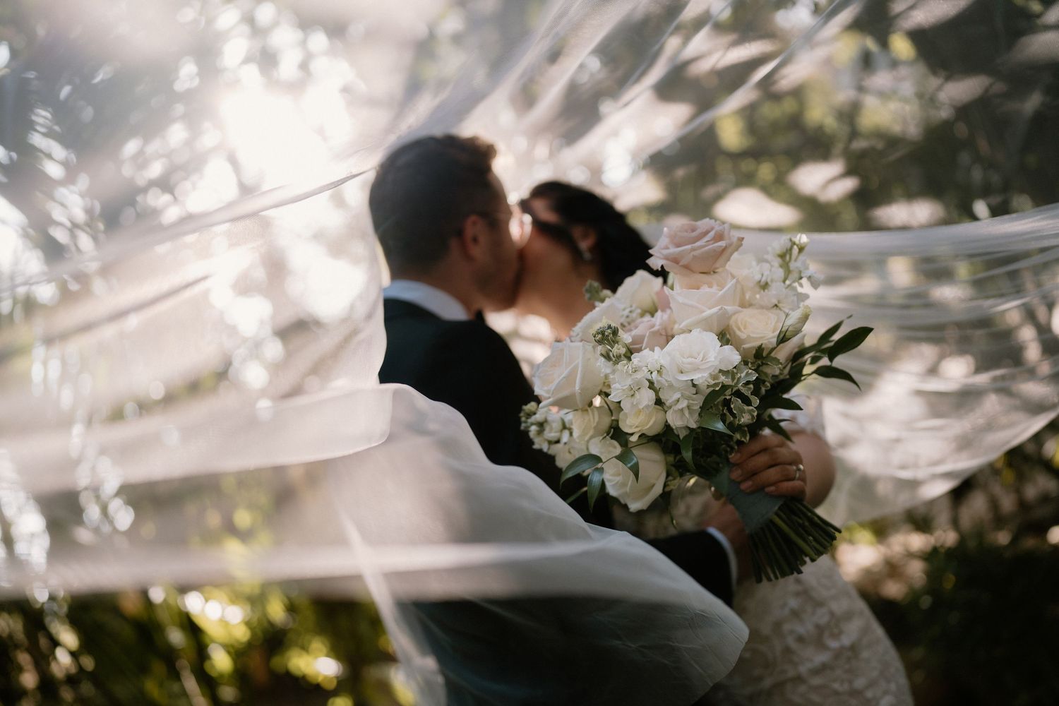 Bride and groom share a romantic kiss under a flowing veil, bride holding white floral bouquet outdoors.