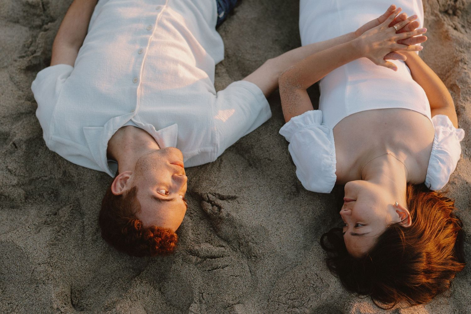 Couple lying on sandy beach laughing together, viewed from above in matching white outfits.