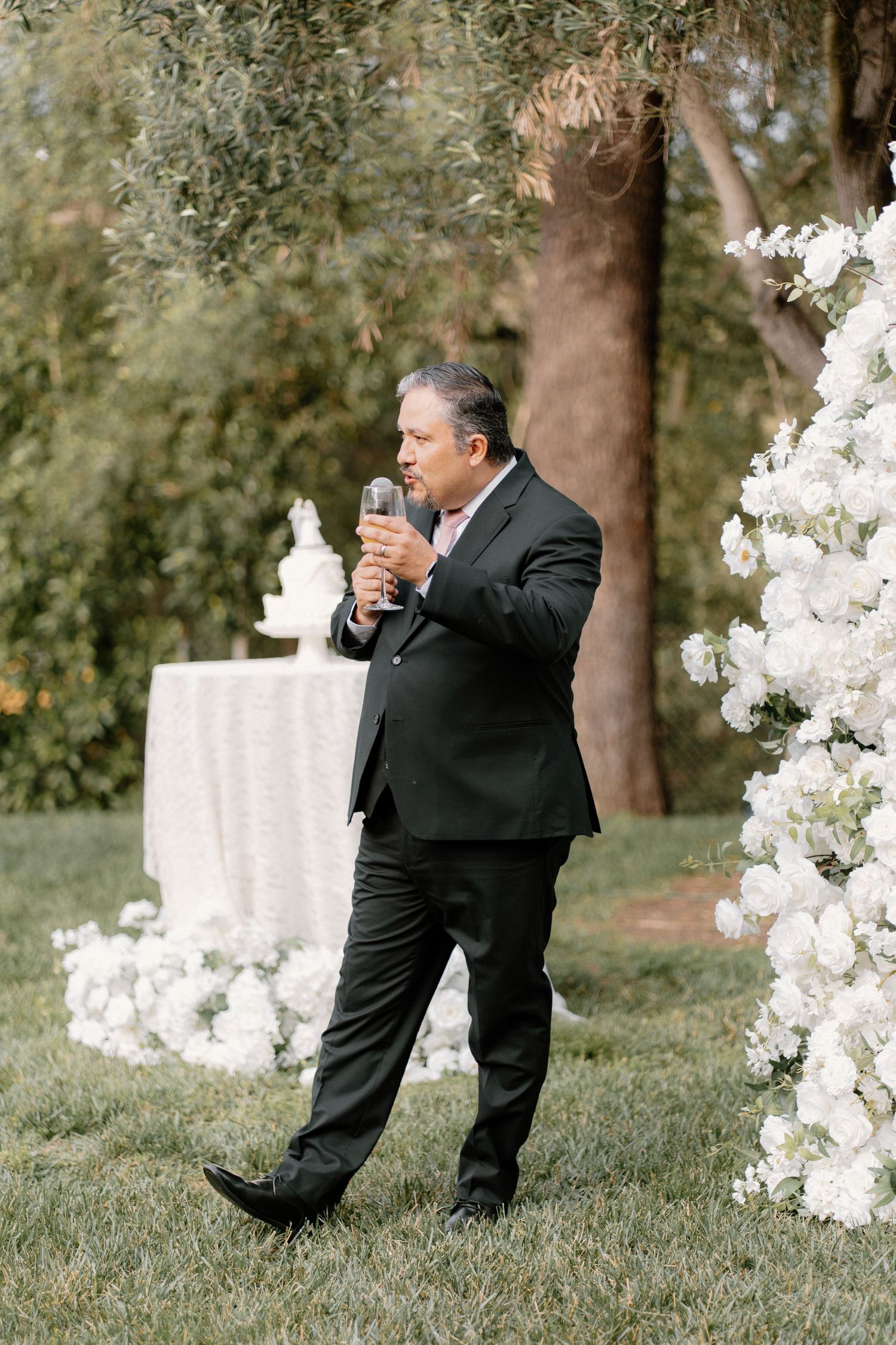 Man in black suit giving a champagne toast at an outdoor wedding ceremony with white floral arch.