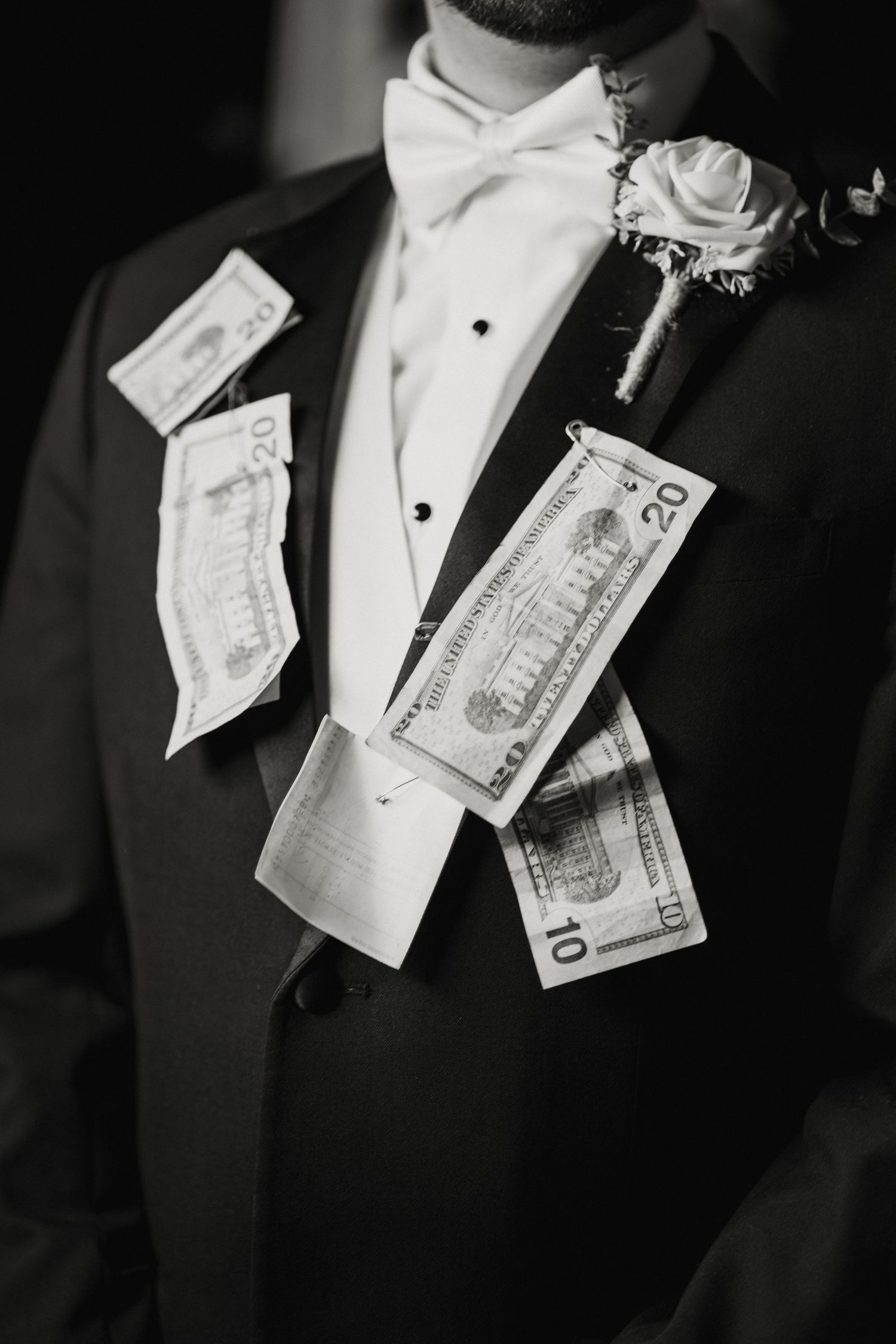 Black and white close-up of groom in tuxedo with dollar bills tucked into jacket at wedding.