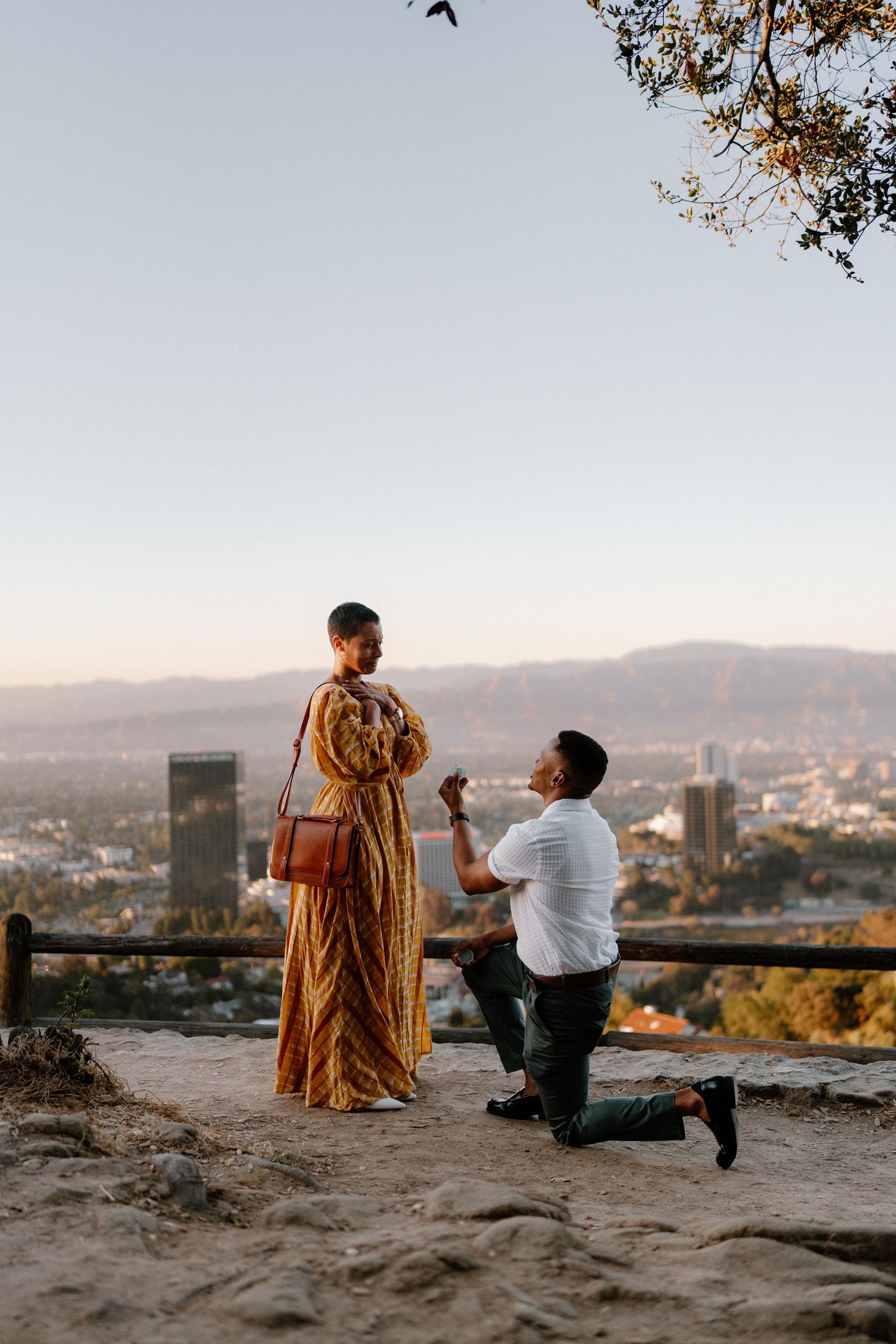A man proposes on one knee to a woman in a yellow maxi dress at a golden hour city overlook.