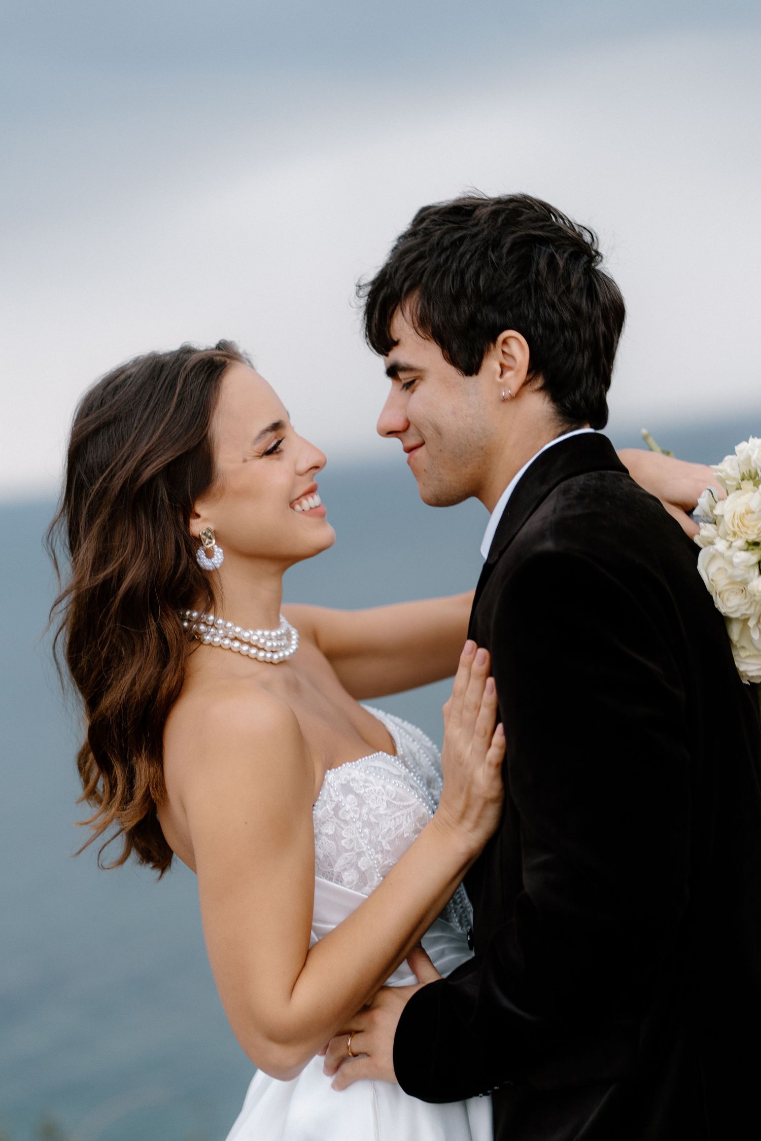Bride and groom gazing at each other near ocean, she wears pearl jewelry and white strapless gown, he holds white bouquet.